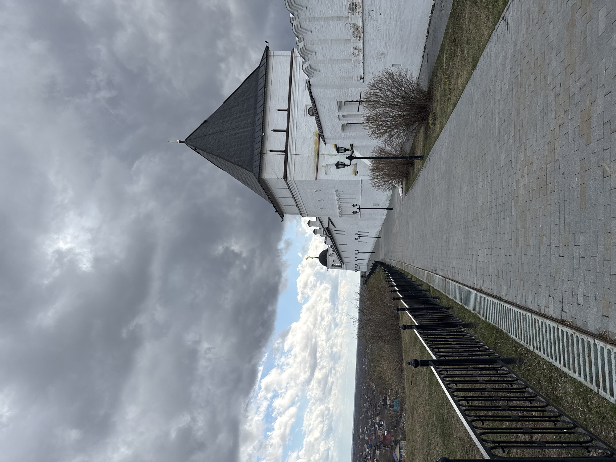 The image captures a section of the Tobolsk Kremlin in Tobolsk, Russia. Specifically, it showcases a portion of the Kremlin wall, constructed from white brick, accompanied by a walkway paved with light gray stones. Adjacent to the walkway is a narrow strip of grassy area where bushes are located, and a black metal railing with decorative posts separates the elevated Kremlin grounds from the cityscape below. The foreground reveals a detailed view of houses and buildings scattered across the landscape. Above, the sky displays a mix of light and dark clouds. The image is rotated 90 degrees counter-clockwise.