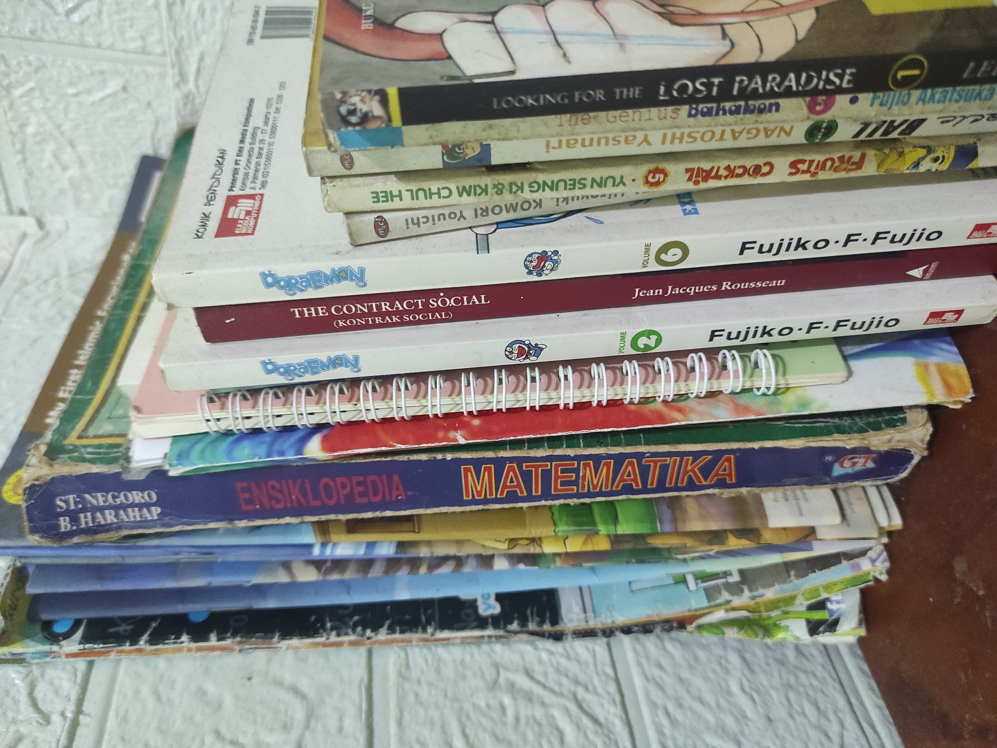 A close-up, high-angle shot shows a stack of books standing upright on a dark wooden surface against a white textured wall. The books are positioned on the left side of the frame, with their spines facing the camera. The spines reveal titles in Indonesian and English, including "ST. NEGORO B. HARAHAP," "ENSIKLOPEDIA MATEMATIKA," "My First Islamic..." and various comic book titles like "THE CONTRACT SOCIAL," "Fujiko F. Fujio," "Fruits Cocktail," "NAGATOSHI YOSHIMURA," and "THE LOST PARADISE." Some of the books appear to be part of a series, indicated by volume numbers and repetitive character illustrations, such as Doraemon. The overall condition of the books suggests they have been used, with some wear and tear visible on the edges and covers. The lighting is even, casting no significant shadows. There is no indication of the specific location in Pleret, Indonesia.