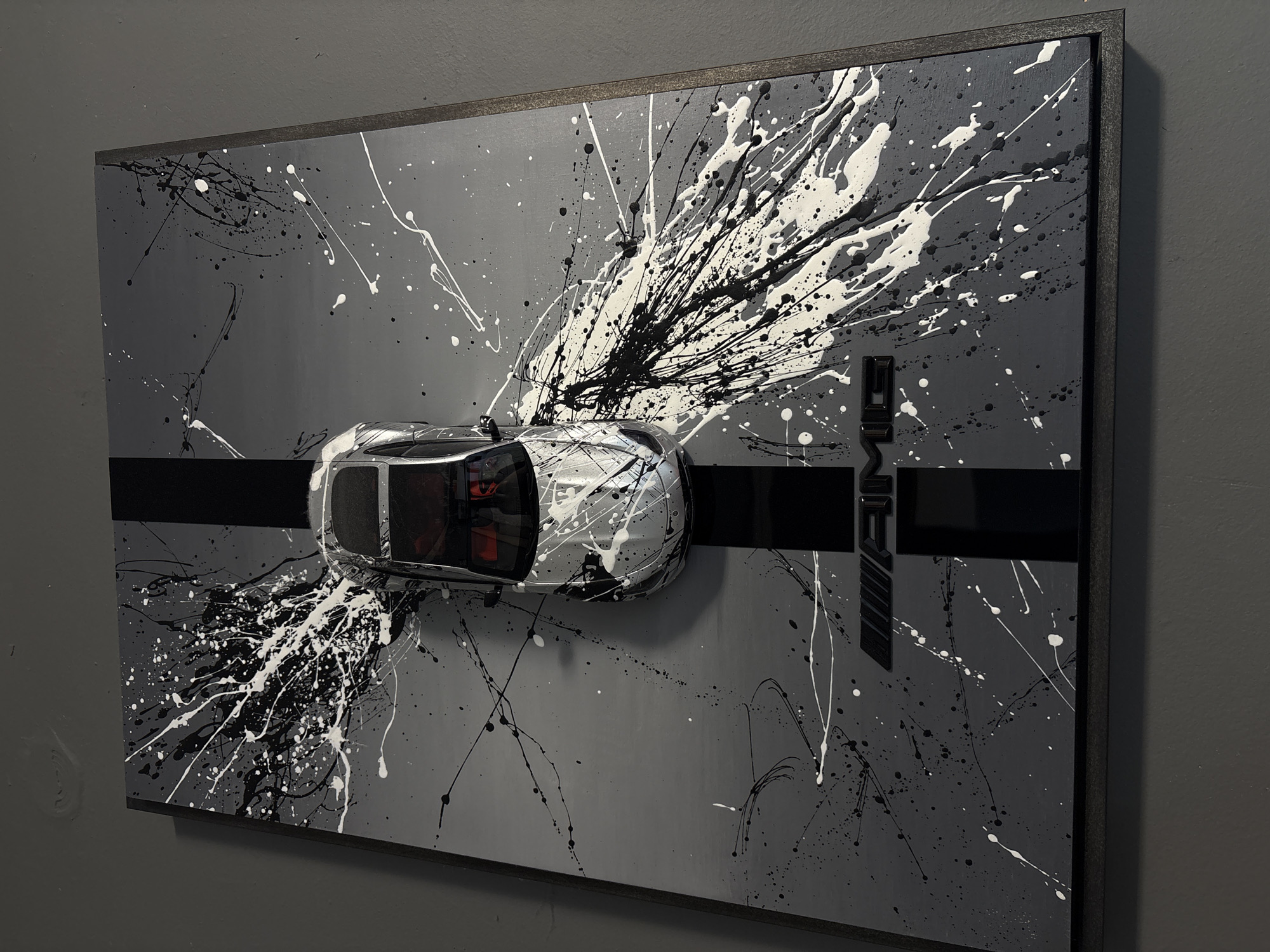 A framed piece of art, possibly a painting or mixed media piece, hangs against a gray wall. The art features a gray background with splatters of white and black paint. A miniature silver-colored car is mounted on the art, with paint splatters also appearing on the car. A thick, black stripe runs horizontally across the center of the artwork. To the right of the car is a black geometric shape or design. The frame of the artwork is dark. There is no indication of the location being Cheshnegirovo, Bulgaria, or any people present in the image.