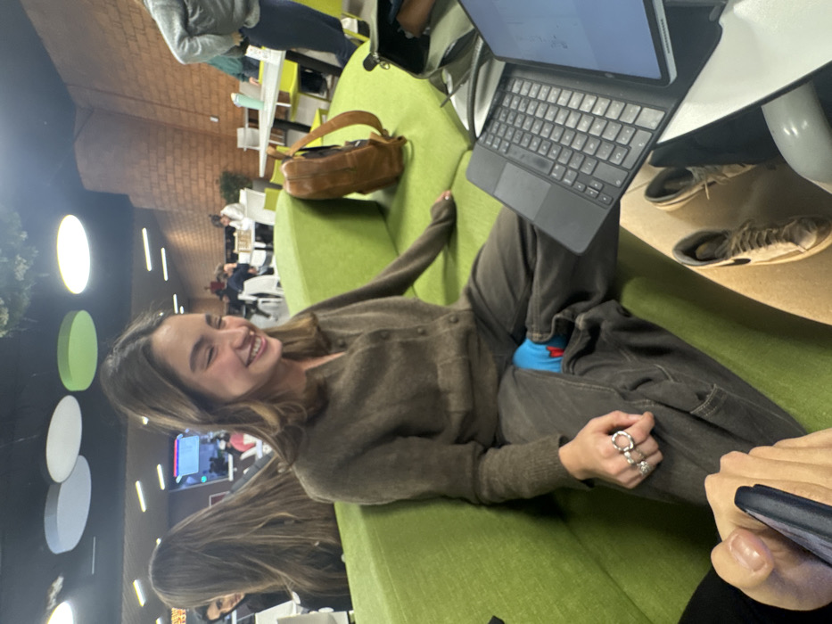 The image portrays an indoor common area, likely a university or public space, situated within Mexico City, Mexico. The precise location is not visually identifiable. In the mid-ground, a young female with medium-length brown hair is seated on a lime-green cushioned sofa, smiling broadly towards the left, outside the frame. She wears a brown button-up cardigan, dark grey trousers, and a visible blue sock with a red pattern on her right foot. Her left hand, adorned with several silver rings, rests on her leg. A black laptop or tablet in a keyboard case is open on an immediate surface or her lap. A brown leather backpack rests on the sofa to her right. In the extreme foreground left, a person's hand holds a dark smartphone. The background reveals multiple individuals seated at white tables with white or lime-green chairs. The ceiling features circular white and lime-green light fixtures, and a reddish-brown brick-patterned wall is visible on the right. A reflection of a person with long brown hair is visible on a dark, vertical reflective surface in the mid-left background.