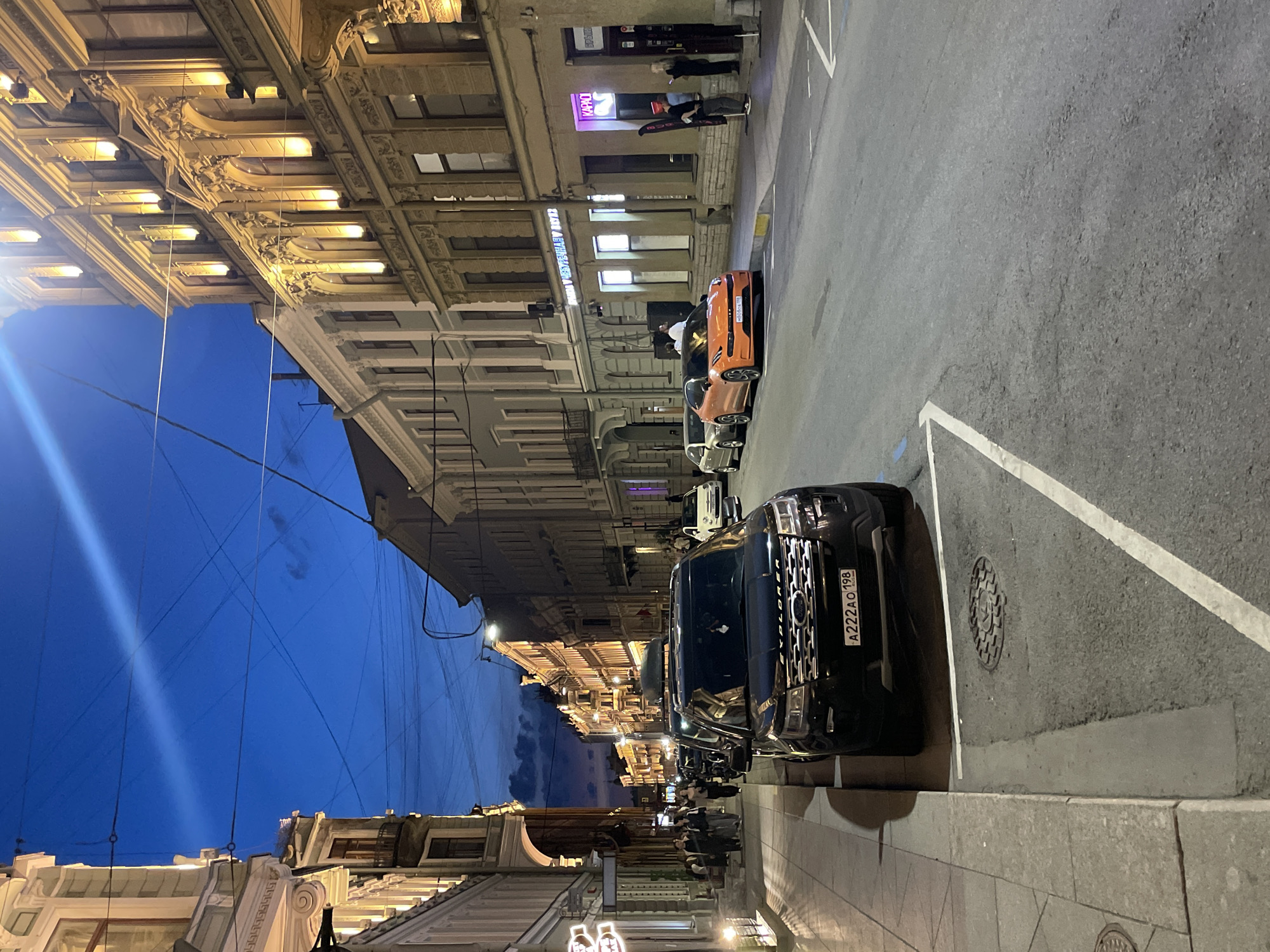 The image depicts a city street scene during twilight or evening, characterized by a deep blue sky and activated artificial lighting. The location is Saint Petersburg, Russia, identifiable by the "198" region code on the license plate of the prominent black Ford Explorer.
Ornate, multi-story buildings with classical architectural elements, including decorative cornices, window frames, and pilasters, line both sides of the asphalt street. Many building facades are illuminated with warm yellow up-lighting. Overhead wires crisscross the sky.
In the immediate foreground on the left, a black Ford Explorer SUV is parked on the sidewalk curb, facing away from the viewer. It bears Russian license plate "A222AO 198" and has a roof rack installed. Further down the street on the right, an orange Lynk & Co 05+ coupe SUV is parked at the curb. Behind it, a silver sedan is visible. Further along the right side, a white compact car is also parked.
On the left sidewalk, a group of approximately five to seven individuals are walking away from the camera. On the right sidewalk, near the orange car, a group of three to four people are standing outside a building with illuminated signs; one sign clearly displays "КАРАОКЕ" (Karaoke) in Cyrillic, and another sign above it partially reads "РЕСТОРАН" (Restaurant). One person in this group wears a distinct red hat. The street itself has clear white lane markings and a yellow curb marking on the right side. Manhole covers are visible on both the street and sidewalk.