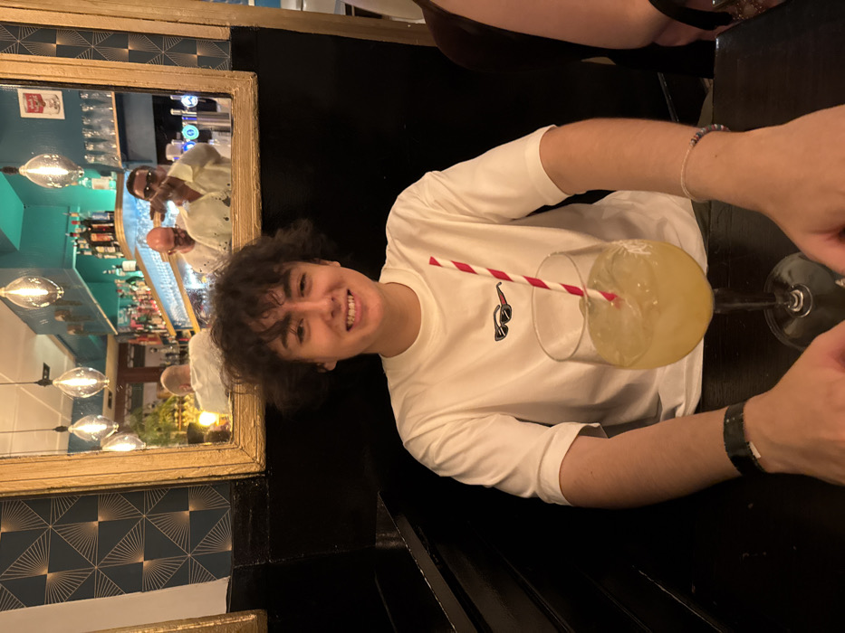 A person smiles beside a cocktail in a tall glass with a red and white striped straw. The individual wears a white long-sleeved shirt. A mirror with a gold frame behind the person reflects a bar area with bartenders and various bottles. The table is black. The person has dark curly hair. A bracelet is visible on the person's wrist.