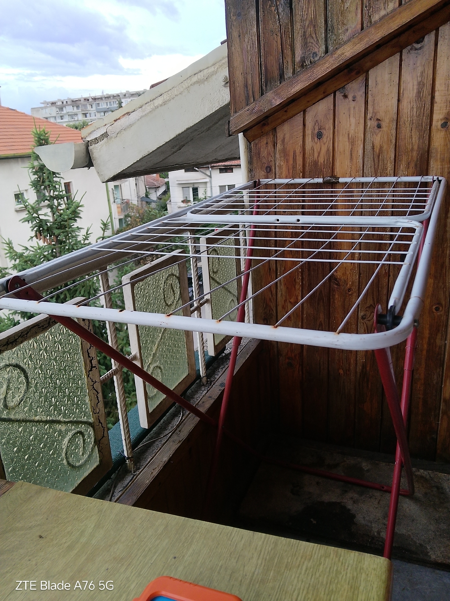 The image captures an outdoor scene, likely a balcony or patio, in Stara Zagora, Bulgaria. In the foreground, a wooden surface with a light yellow hue is visible, featuring the text "ZTE Blade A76 5G". Dominating the center is a white clothes drying rack with red legs. The drying rack has multiple horizontal bars and diagonal supports. Behind the drying rack, a wooden wall composed of vertical planks shows signs of weathering and discoloration. To the left of the drying rack, a balcony railing with decorative, textured green glass panels can be seen. In the background, a residential building with a red-tiled roof and another taller, modern building with multiple floors are visible under a cloudy sky. A fir tree with pinecones is also present. The ground beneath the drying rack appears to be concrete and is damp in some areas. The overall impression is one of a functional outdoor space, possibly aged, with residential buildings and natural elements in the backdrop.