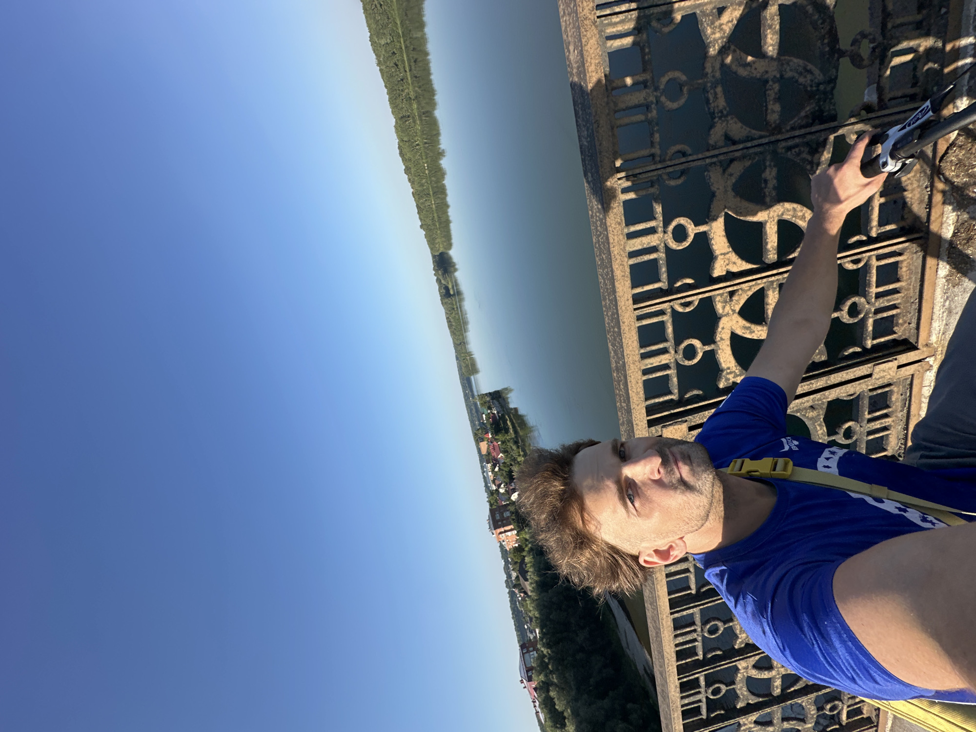 A man with tousled brown hair, wearing a blue t-shirt and a yellow strap across his chest, is taking a selfie on a bridge. He is positioned in the foreground, with a wide river and a tree-lined bank stretching into the distance behind him. A town with red-roofed buildings is visible on the left bank of the river. The bridge railing, with its ornate metalwork, is also a prominent feature in the foreground. The sky is clear and blue. This scene appears to be on the Yablonitsky Bridge over the Biya River in Biysk, Russia, given the characteristic architecture of the town and the natural landscape.