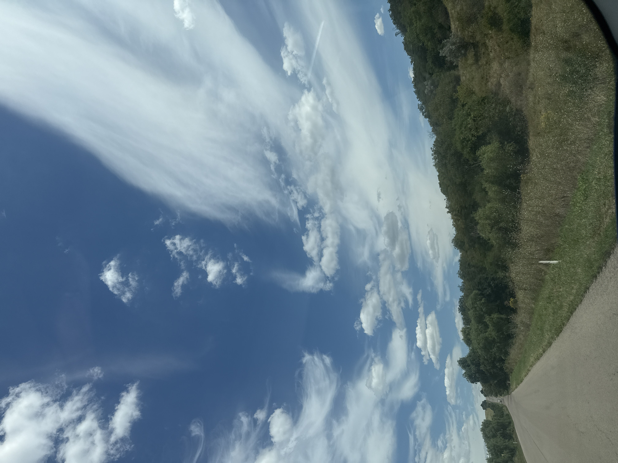 An eye-level, medium outdoor shot shows a paved road winding uphill through a landscape of trees and vegetation. The sky is a vibrant blue, with white, wispy clouds scattered across it, some resembling streaks. The road is empty in the foreground and recedes into the distance where a white vehicle is visible. To the right of the road, a grassy embankment is covered with dry grass and a few small white markers. A dense line of green trees covers the hillside to the right of the road, extending into the background. The overall impression is one of a rural, scenic road on a bright, partly cloudy day, potentially in or around the city of Iurievca, Moldova.
