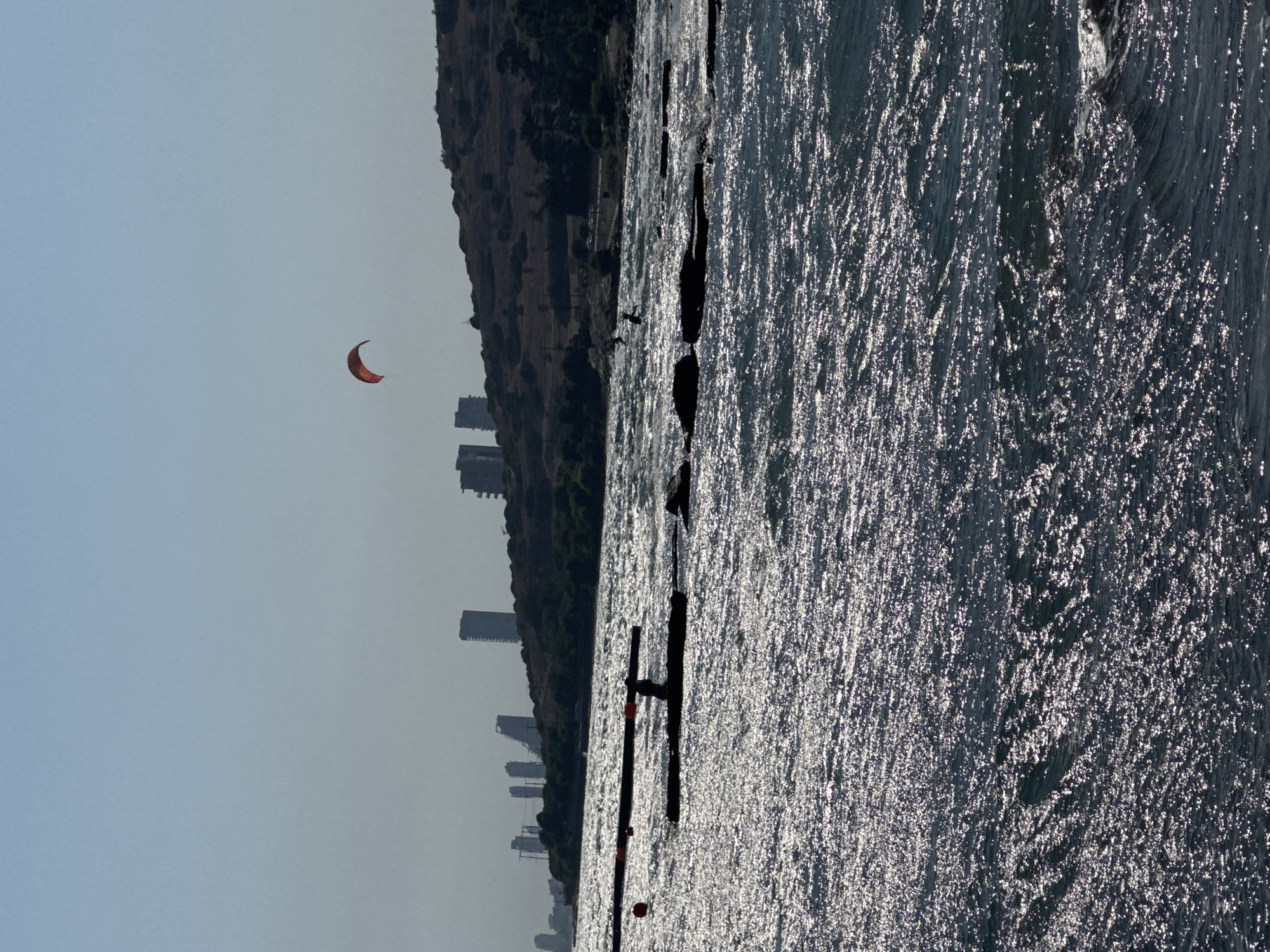 The image shows a bright, sunny day at the sea with a clear blue sky. In the foreground, the dark blue water is choppy with sunlight glinting off the surface. A person is visible on a dark, flat structure in the water, and another person is seen further in the distance. Two red buoys are floating near the structure. In the background, a line of buildings, some tall and modern, are silhouetted against a dry, hilly landscape. A small, orange kite is visible in the sky above the hills. The overall scene suggests a recreational area by the sea, likely a beach or a bay within the Agios Tychona Community, Cyprus, given the modern architecture and coastal setting.