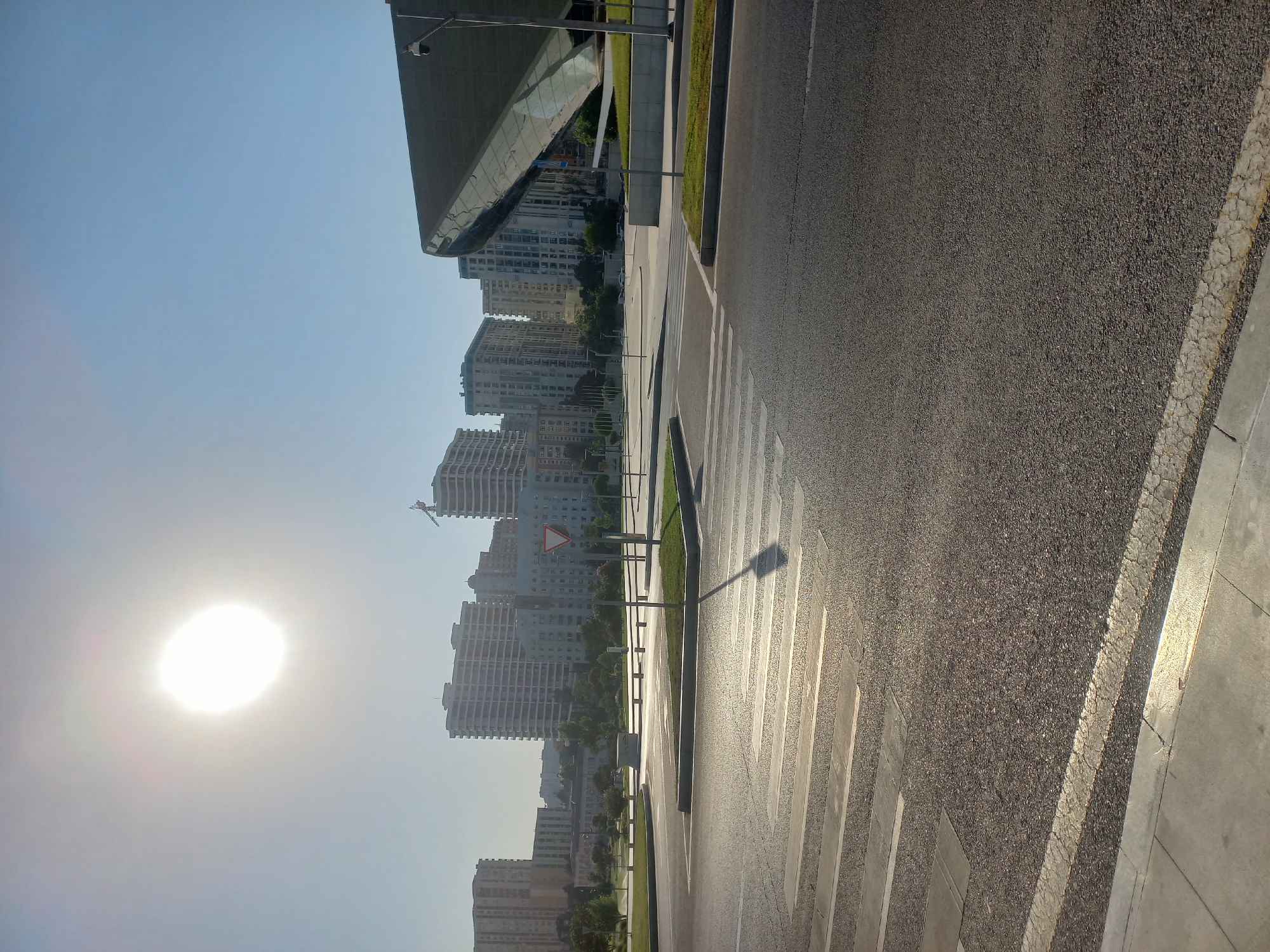 The image captures an exterior view of the Baku Congress Center and surrounding area in Baku, Azerbaijan, under a bright, sunny sky. The sun is high and slightly to the left, casting a strong glare and creating distinct shadows. A wide asphalt road dominates the foreground, featuring a marked pedestrian crossing. To the right, the distinctive, futuristic architecture of the Baku Congress Center, with its large, reflective, angled roof, is visible. In the background, several multi-story apartment buildings and a construction crane suggest a developing urban landscape. The environment appears clean and well-maintained, with manicured green spaces and streetlights visible along the road. The scene is devoid of any people or vehicles, giving it a tranquil and almost staged appearance.