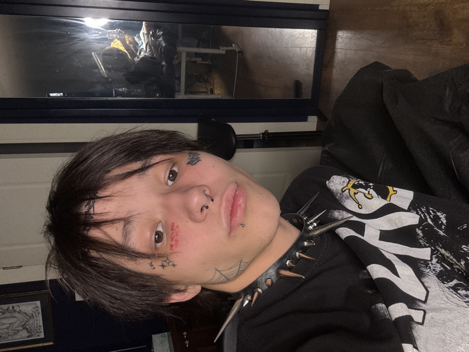 A person with dark hair and facial tattoos is angled towards the camera. The tattoos include characters and a phrase on the face. The person has lip and nose piercings, and wears a black spiked collar. Part of a graphic black and white shirt is visible. A dark-framed mirror shows a cluttered reflection.