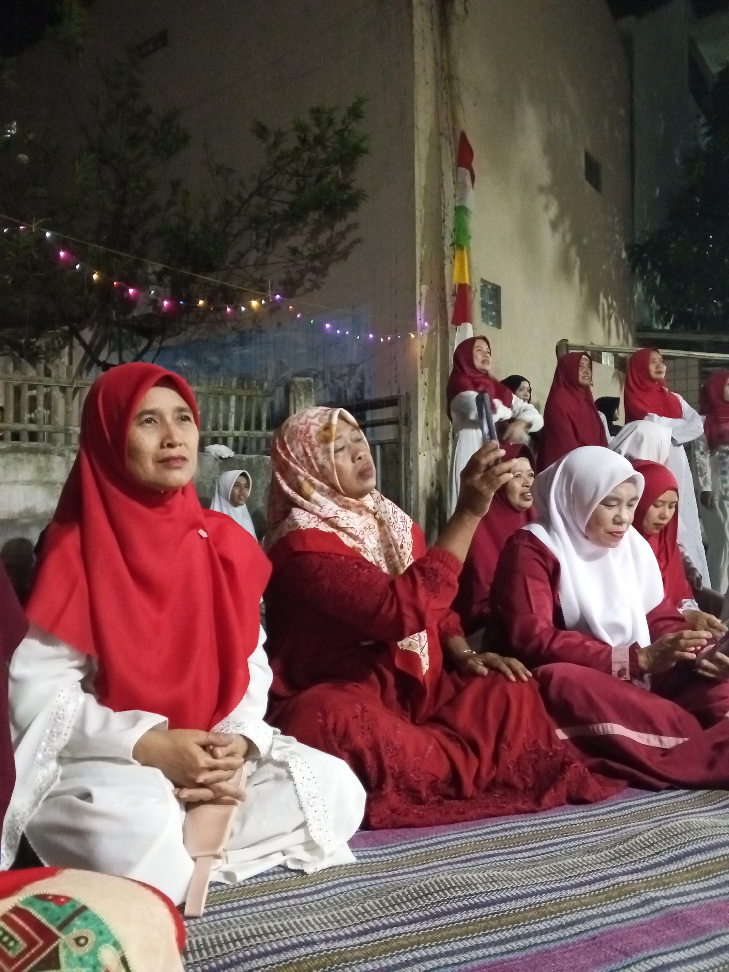 The image depicts a gathering of women, many wearing red and white hijabs, seated on the ground. The scene appears to be outdoors at dusk or night, indicated by artificial lighting in the background and the dark sky. The women are dressed in traditional Islamic attire, with some in long dresses and headscarves. The setting suggests a community event or religious observance. A wall and some fencing are visible in the background, along with trees and some decorative lights strung across an area. The ground is covered with a woven mat. The presence of a flag or banner with red and white stripes on the wall may indicate a patriotic or cultural event. Based on the context provided about the location, this scene is situated in Ciruas, Indonesia.
