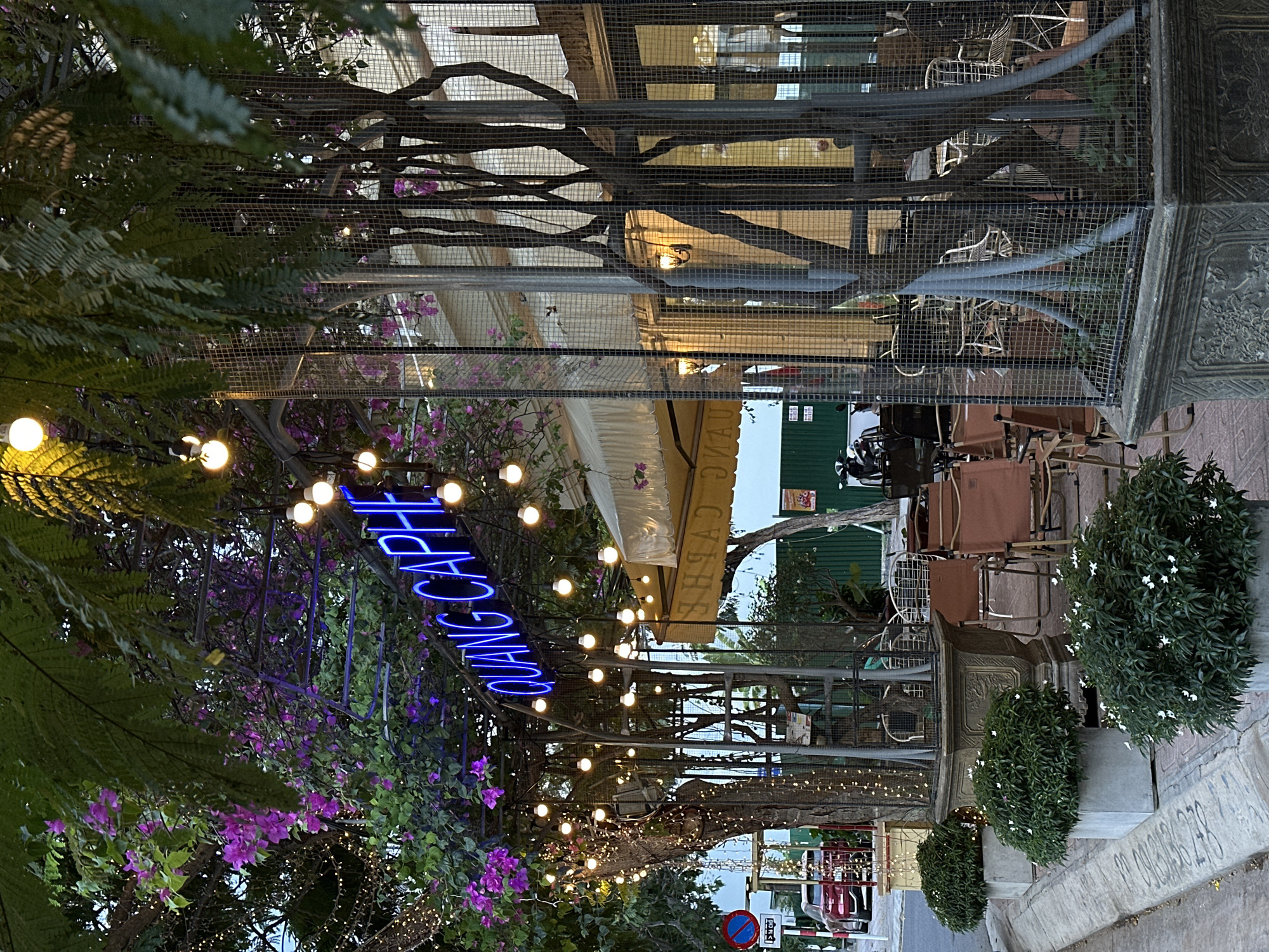 The image depicts an outdoor seating area of a cafe in Nam Nha Trang Ward, Vietnam. The scene is adorned with abundant greenery, including ferns and flowering plants with purple blossoms, interspersed with string lights that illuminate the space. A prominent neon sign in blue, spelling "QUANG CAPHE," is visible, indicating the cafe's name.
The cafe's ambiance suggests it is evening or dusk, with the artificial lights creating a warm and inviting atmosphere. The seating area features tables and chairs, some made of rattan and others with brown upholstery, set against a backdrop of trees and wire mesh fencing. Architectural elements, including ornate metalwork and a decorative pillar, add to the cafe's character.
In the background, a glimpse of a street can be seen, with a motorbike parked and some buildings suggesting an urban setting. The weather appears to be clear, suitable for outdoor dining. The overall impression is one of a lively and pleasant gathering spot, likely popular for its atmosphere and the aesthetic appeal of its decor. No specific activities or people are clearly discernible, but the setup implies a place for relaxation and socializing. Visible text includes the cafe's name "QUANG CAPHE" on the neon sign and some unreadable text on a sign in the background.