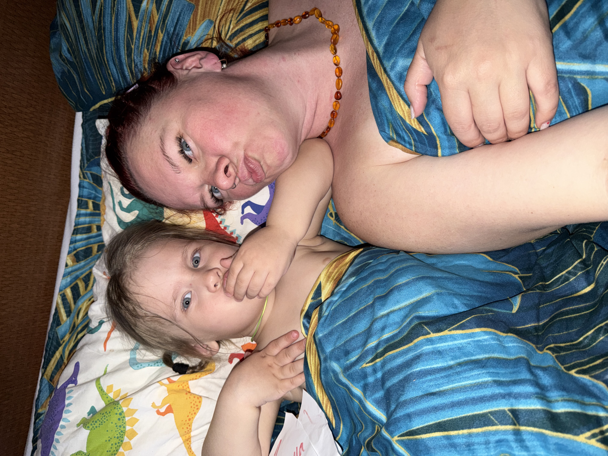 A woman and a young child are lying on a bed. The woman, wearing an amber necklace, is positioned at the top of the image with a playful expression. The child, with blonde hair, is lying below her, partly covered by a blue and gold patterned blanket. The bed has a dinosaur-themed sheet. The scene is indoors. Location: Likely Chaikovsky, Russia.