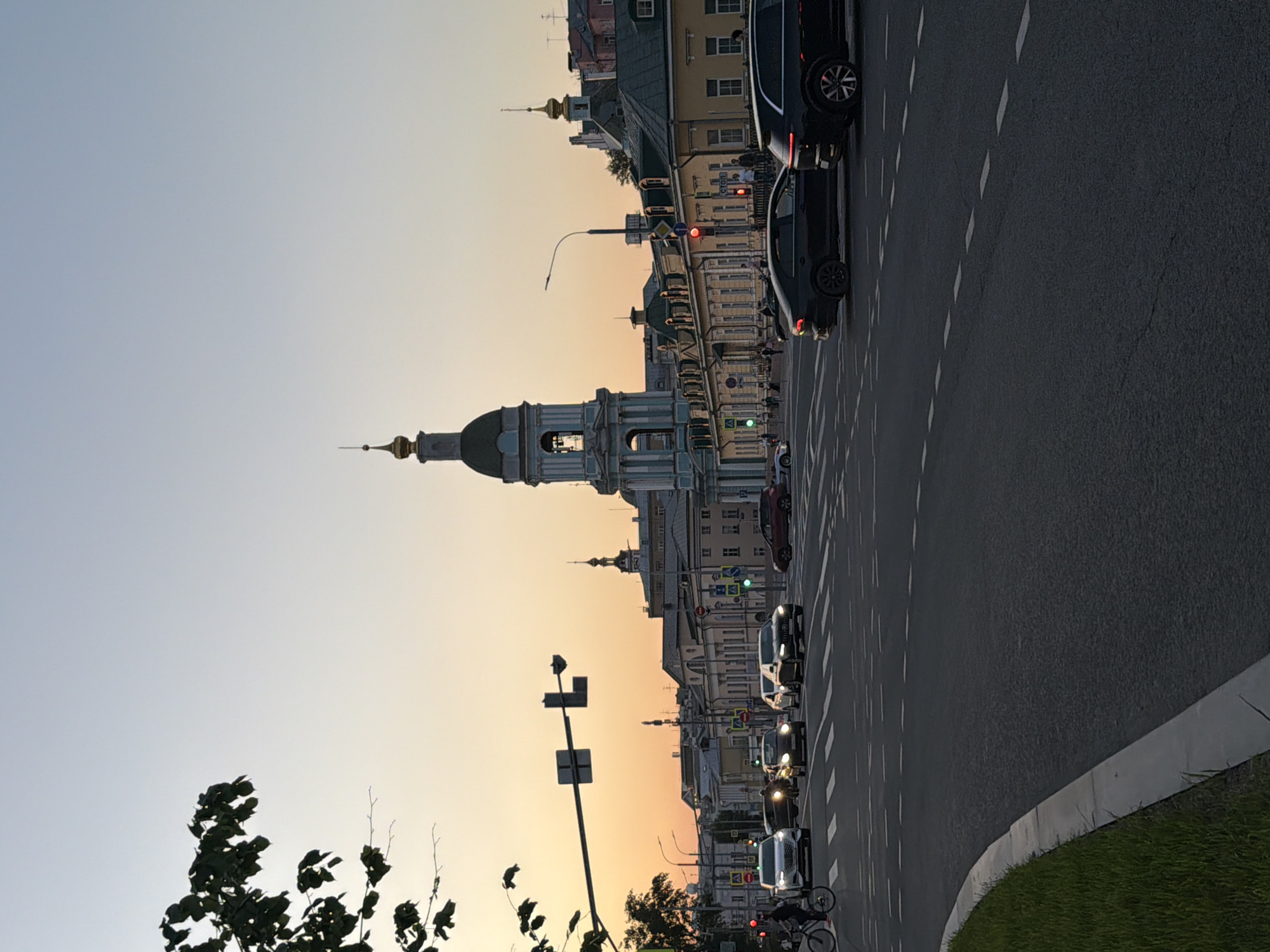 The image depicts a street scene in Moscow, Russia, at dusk. The sky transitions from a pale blue to a warm orange, suggesting either sunset or sunrise. A prominent architectural feature is a church or cathedral with a green dome and a tall bell tower. Several buildings line the street, varying in height and architectural style. A row of vehicles, including cars and what appears to be a motorcycle, are positioned on the right side of the frame, on a multi-lane asphalt road marked with dashed white lines. A pedestrian is riding a bicycle on the right side of the road. Green grass borders the edge of the roadway. Traffic lights are visible, with one showing a red light.