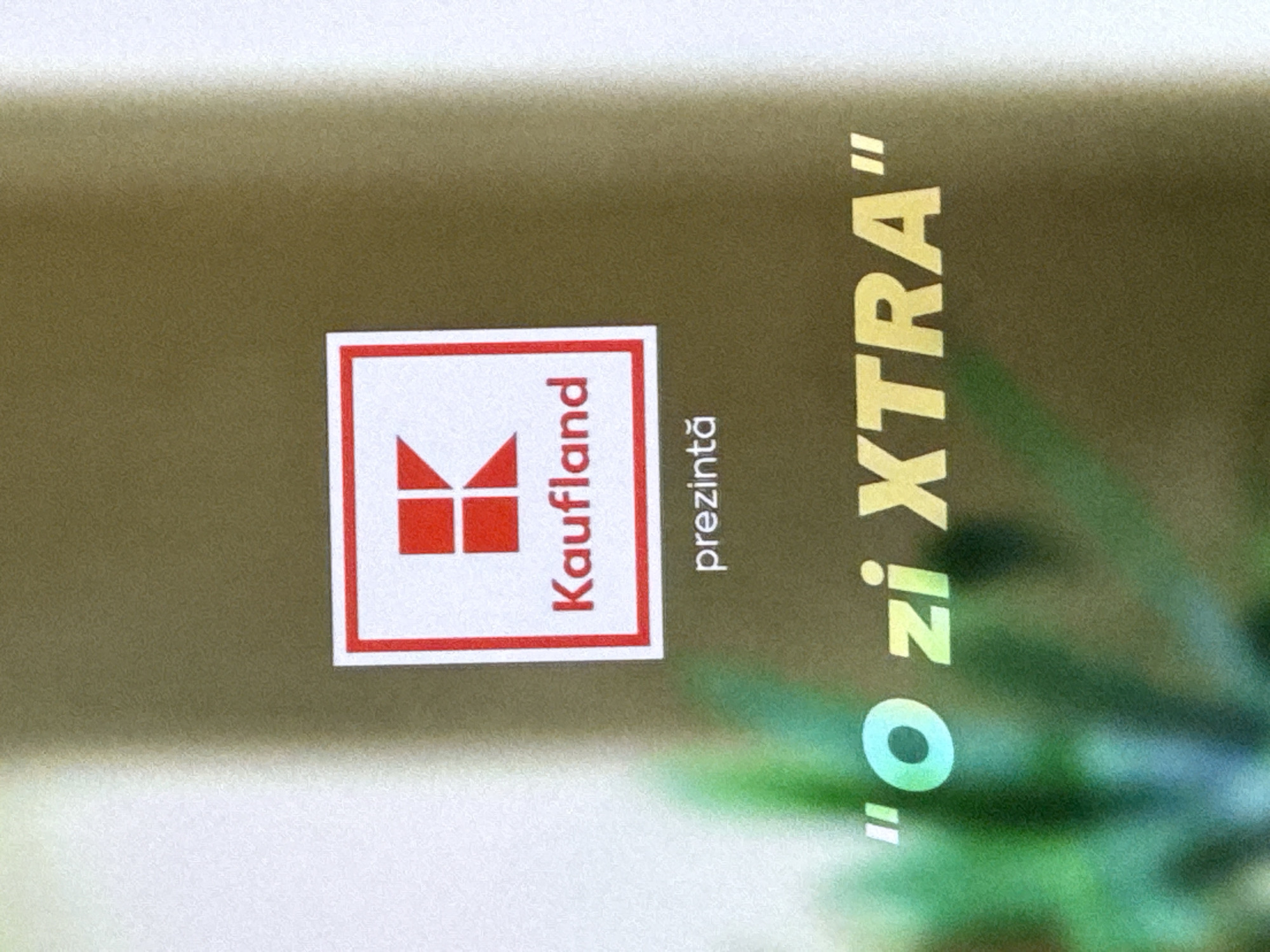 The image shows a close-up of a sign for Kaufland, a supermarket chain, in Cahul, Moldova. The Kaufland logo, a red "K" on a white background within a red square border, is visible in the upper center of the image. Below the logo, the word "Kaufland" is printed in red. Further down, the word "prezintă" is written in smaller black text. At the bottom of the image, a slogan in large yellow letters reads "O zi XTRA". The background is out of focus, showing a gradient of colors that appear to be a light brown and green, possibly from the store's interior or exterior surroundings. There are no people or discernible actions depicted in the image, only the branding and slogan.