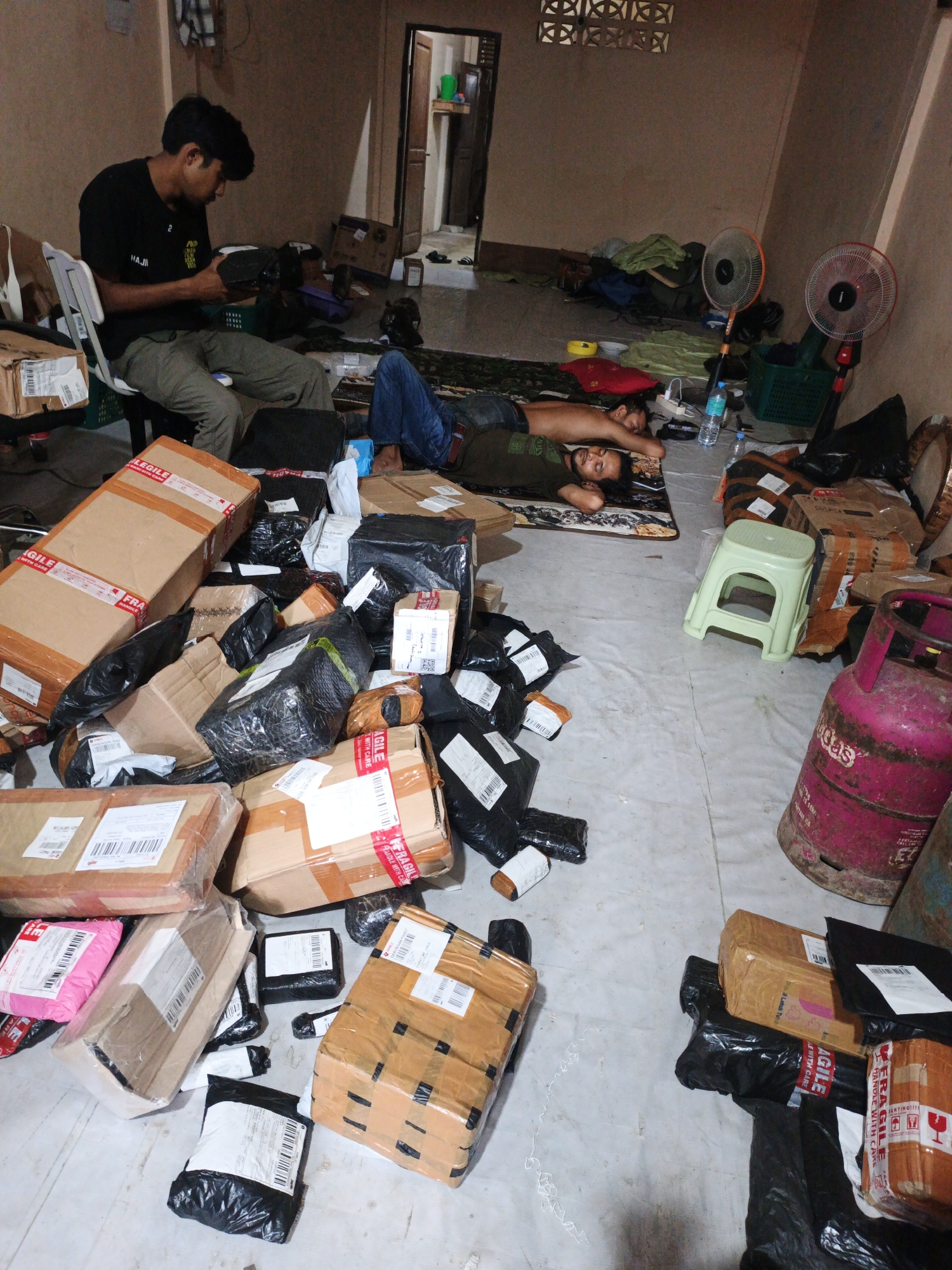 The image depicts an indoor scene within what appears to be a small, cluttered room in Lhokseumawe, Indonesia. The floor is covered with numerous packages of various sizes, many wrapped in brown cardboard and sealed with tape, others in black plastic. Some packages have shipping labels and "FRAGILE" tape. Two men are present in the room. One man is seated on a white chair, wearing a black t-shirt and khaki pants, and is holding and examining a black package. The second man is lying down on the floor, appearing to be asleep, with his head resting on a pillow and his body covered by a dark green t-shirt and blue jeans.
In the background, there are two electric fans, one orange and one red, both positioned on the floor. A green plastic basket is visible, containing some items. A pink propane tank is prominent on the right side of the image. The walls of the room are a muted brown color, and there is a doorway visible in the far left, leading to another area. The overall impression is one of a room used for storage and possibly shipping, with the occupants resting amidst the clutter. The lighting suggests it is daytime, but the room is not well-lit.