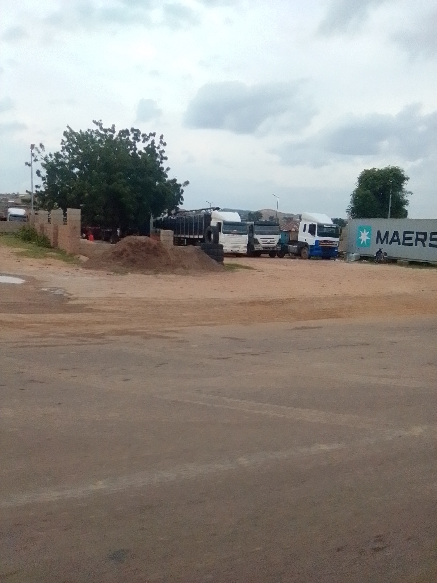 A wide shot captures a lot with multiple trucks parked under an overcast sky in Gombe, Nigeria. The foreground is a dusty, light brown road leading towards a lighter brown dirt lot filled with construction materials such as sand and bricks. Behind the lot, several white semi-trucks are parked. To the right of the trucks, a large white shipping container with the Maersk logo is visible. In the background, lush green trees and a few small buildings are scattered across the landscape. A motorcycle with a rider is parked near the shipping container. The sky is filled with gray clouds, indicating a possibility of rain.