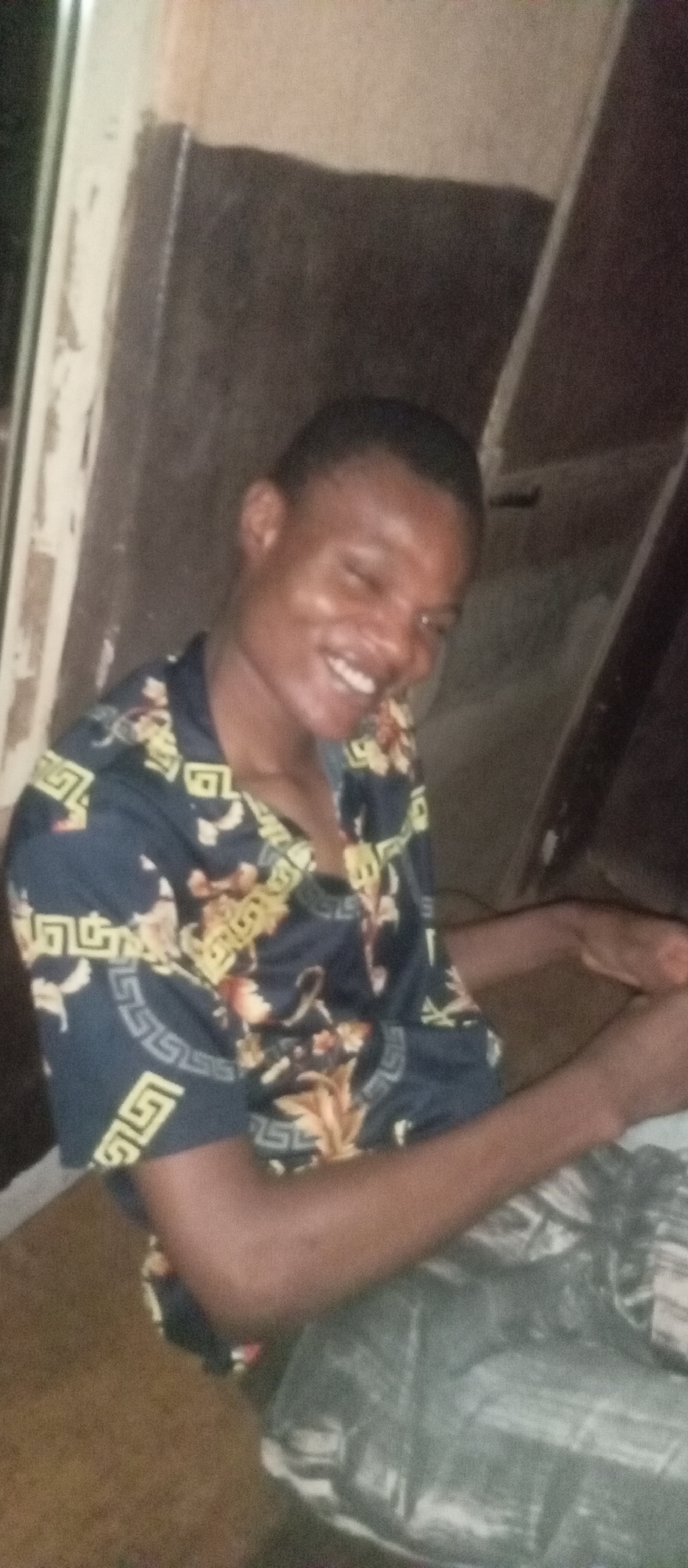 A young male, appearing to be in his late teens or early twenties, is depicted in a medium close-up, smiling broadly at the camera. He has short, dark hair and a cheerful expression, with his teeth visible in a wide smile and eyes squinted in happiness. He is wearing a dark, possibly navy blue, collared shirt featuring an intricate pattern of golden-yellow floral or leafy designs, interspersed with geometric Greek key (meander) patterns along the sleeves and borders. His right arm is visible, bent at the elbow.
The setting appears to be indoors and dimly lit, suggesting it might be evening or night. The background consists of dark, likely wooden or painted walls, with a lighter vertical element visible on the left side, possibly a doorframe or part of a structure. The overall impression is one of casual comfort and happiness. This image was taken in Ogidi, Nigeria.