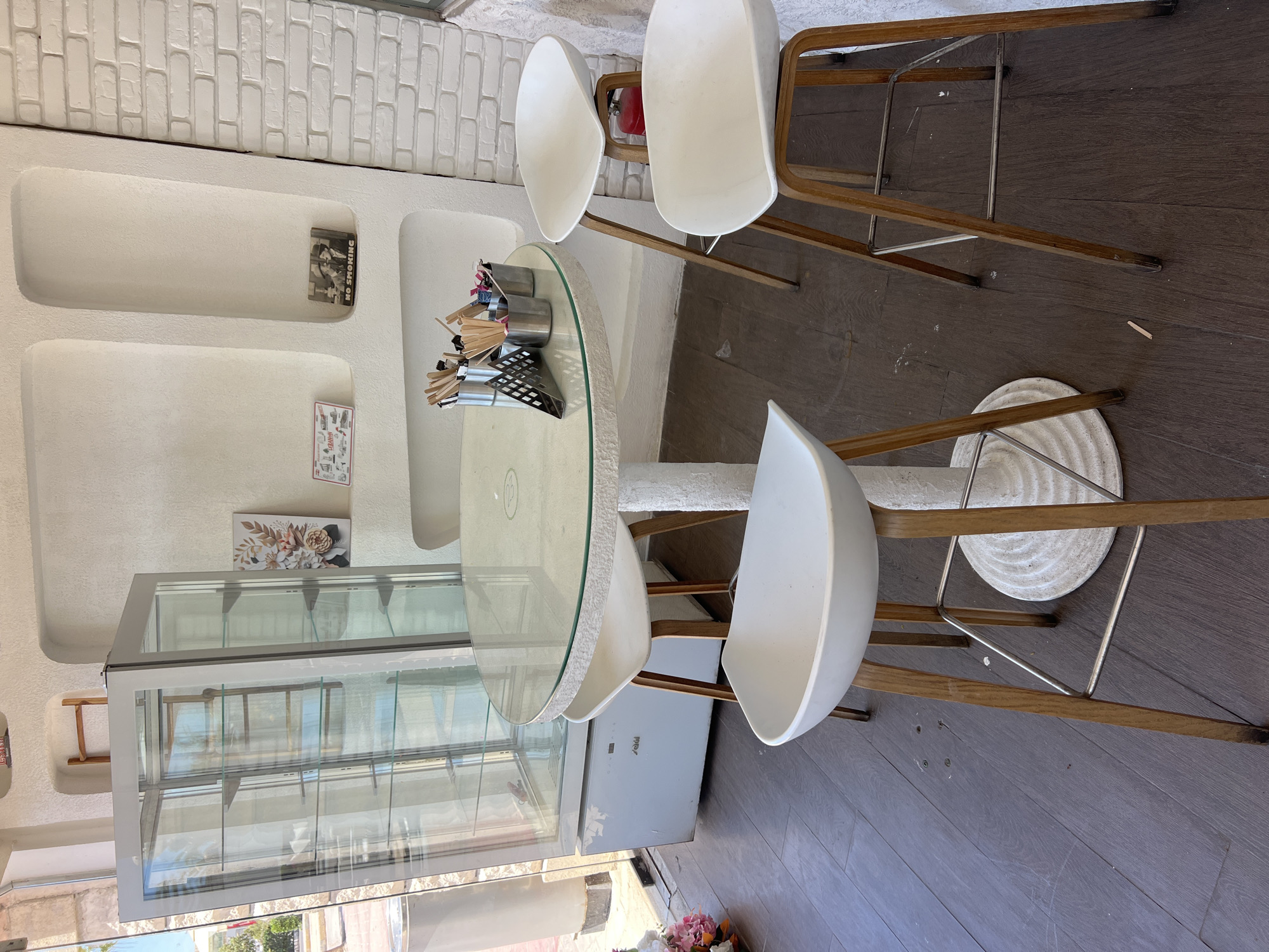 This is an indoor scene featuring a small cafe or restaurant seating area. The subjects are primarily furniture and decorative items. There is a round glass-top table with a textured white rim, surrounded by three white modern chairs with wooden legs. Two of the chairs are bar stools. The table has a metal napkin holder and several metal containers filled with what appear to be stirrers or cutlery.
Behind the table, there's a display case with glass doors and shelves, suggesting it's used for showcasing food or merchandise. The wall behind the furniture has built-in niches, one of which holds a framed picture and a "NO SMOKING" sign. Another niche displays a floral artwork. The floor is made of dark wood planks. The overall aesthetic is clean and modern, with white walls and contrasting dark flooring.
The lighting suggests it is daytime, with natural light entering from the right side of the frame, casting shadows. The presence of the display case and seating implies that this is a commercial establishment, likely a cafe or dessert shop, possibly in Sharm El Sheikh, Egypt, as indicated by the location context. There is no visible activity or people in the image. Notable details include the specific design of the chairs, the items on the table, and the decorative elements on the wall. Text visible includes "NO SMOKING" and some unreadable text on a small sign on the wall, and "VIRA" on a light grey appliance.