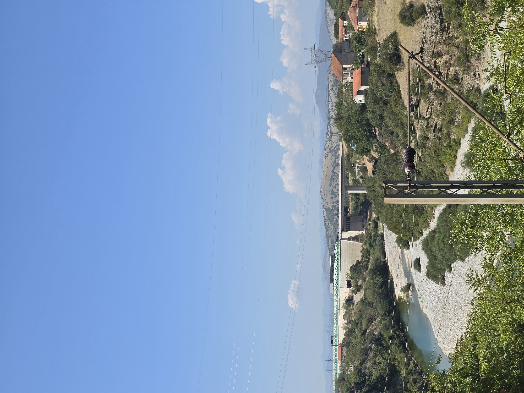 A wide, sunlit, vertical shot shows a clear blue sky dominating the upper two-thirds of the frame. Below the sky, a wide, shallow river with a pebble-strewn bed curves from the left towards the center of the image. The river is bordered by lush green vegetation and rocky terrain.
In the background, a large, imposing concrete dam stretches across the horizon. A bridge extends from the dam, supported by thick concrete pillars. Beyond the dam and bridge, rolling hills recede into the distance, some covered in sparse vegetation and others appearing barren and rocky. A few scattered houses with red-tiled roofs are nestled on the slopes of the hills. A tall, lattice-like power pylon stands to the right of the houses, with power lines stretching across the frame.
In the foreground, on the right side, a metal pole with electrical insulators and wires is prominently visible, suggesting the presence of a power line infrastructure. The overall impression is of a rural or semi-rural landscape with significant infrastructure. The scene is bright and clear, indicative of a sunny day. The location is likely near the Morača River in Podgorica, Montenegro, given the characteristic landscape and infrastructure elements like the dam and power lines, which are common in the area. There are no people visible in the image.