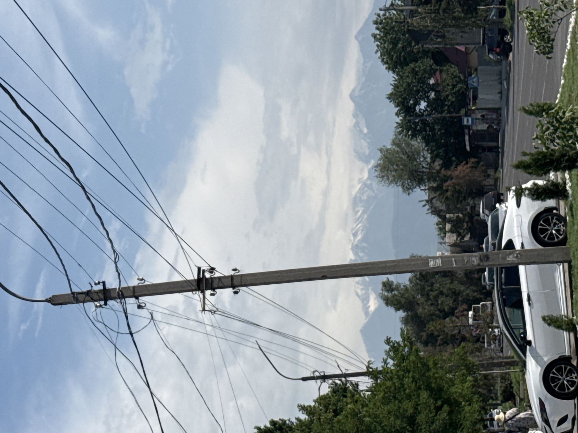 An outdoor, medium shot shows a street scene with a prominent utility pole and a white car in the foreground. The utility pole, made of concrete, stands to the right of the frame, adorned with numerous black electrical wires that crisscross the sky. Behind the pole, a street lamp on another pole is visible.
A white sedan with a black roof is parked on the side of the road, facing left. A person wearing a light-colored hat and a patterned shirt stands near the car on the sidewalk, partially obscured by a green tree.
In the background, a line of trees separates the street from a residential area with houses featuring red roofs. Beyond the trees, a mountain range with snow-capped peaks stretches across the horizon under a cloudy sky. The sky is a pale blue with scattered white clouds. The overall scene depicts an urban environment with a natural backdrop.