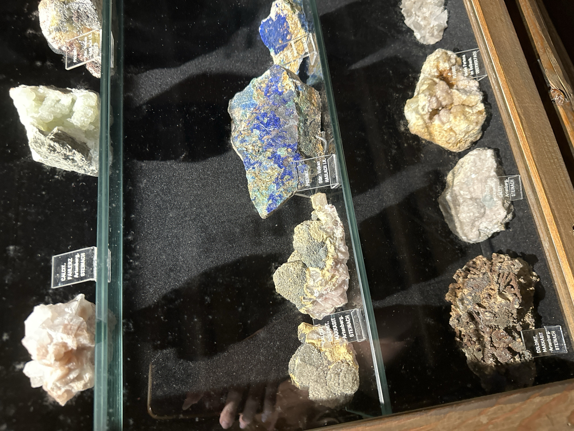 This media file captures a close-up view of a display case filled with various mineral specimens. The minerals are presented on a dark, textured surface, likely velvet, and illuminated from above. Each specimen is accompanied by a small, transparent label detailing its type and origin, such as "CALCIT, FAHLERZ Artenberg, STEINACH" and "FLUORIT Grube Fortuna Im Belbach, OBERWOLFACH."
The collection features a variety of rock types and colors, including pale green, blue, white, and earthy brown specimens. One prominent specimen in the center is a large piece of rock with striking blue and golden-brown veins, identified with labels like "PREHNIT Stbr. Vulkan HASLACH I.K." Other notable minerals include a cluster of pale pink, crystalline formations and a rough, dark brown mineral specimen. The display is housed within a wooden frame, suggesting it is part of a museum, collection, or shop. The lighting creates shadows and highlights, emphasizing the textures and facets of the minerals. The overall impression is one of a well-curated geological display. The provided location context of Schonach im Schwarzwald, Germany, suggests this is a local collection or exhibit of minerals from Germany.