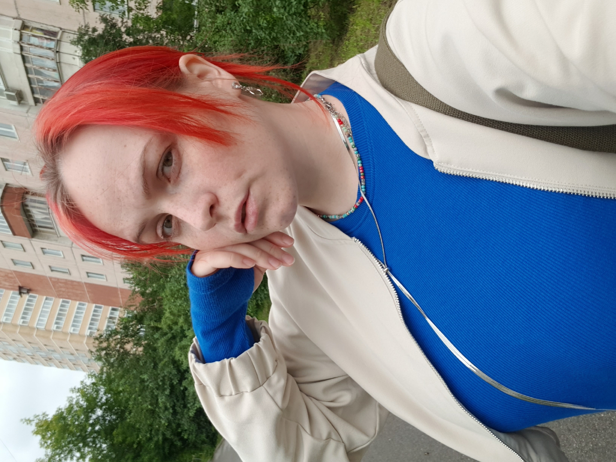 A medium shot is captured from a slightly low angle of a young woman with bright red hair, freckled skin, and light-colored eyes. She is looking directly at the camera with a neutral expression, her right hand resting on her cheek. She is wearing a bright blue long-sleeved shirt and an off-white bomber jacket. A beaded necklace in various colors is visible around her neck, along with a thin silver chain. A khaki strap from a bag crosses her shoulder. The background features a multi-story apartment building and lush green trees. The scene appears to be outdoors in an urban environment.