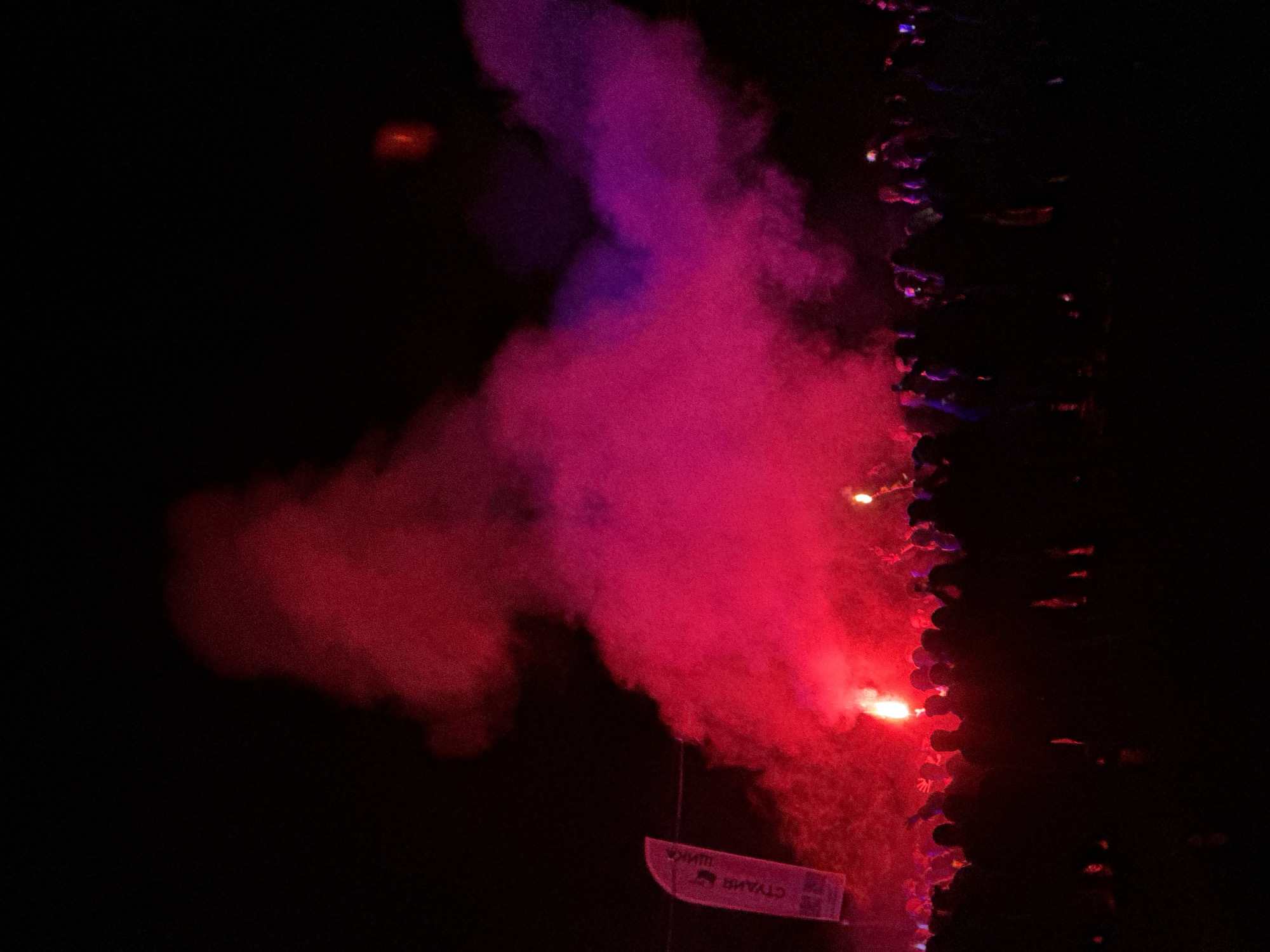 The image depicts a nighttime outdoor gathering in Большая Кивара, Russia. A dense crowd of people, largely silhouetted, occupies the lower third of the frame. Their forms are indistinct, though some individuals appear to have arms raised. Multiple bright flares, emitting intense red light and voluminous red smoke, are visible within the crowd and rising above it. The red smoke dominates the middle ground, expanding upwards and blending with some purple-hued smoke on the right side of the frame. On the far left, a white, vertical banner with Cyrillic text, including "СТУДИЯ", is partially discernible against the dark background. The environment is dark, illuminated primarily by the flares and the colored smoke, indicating a nighttime event.