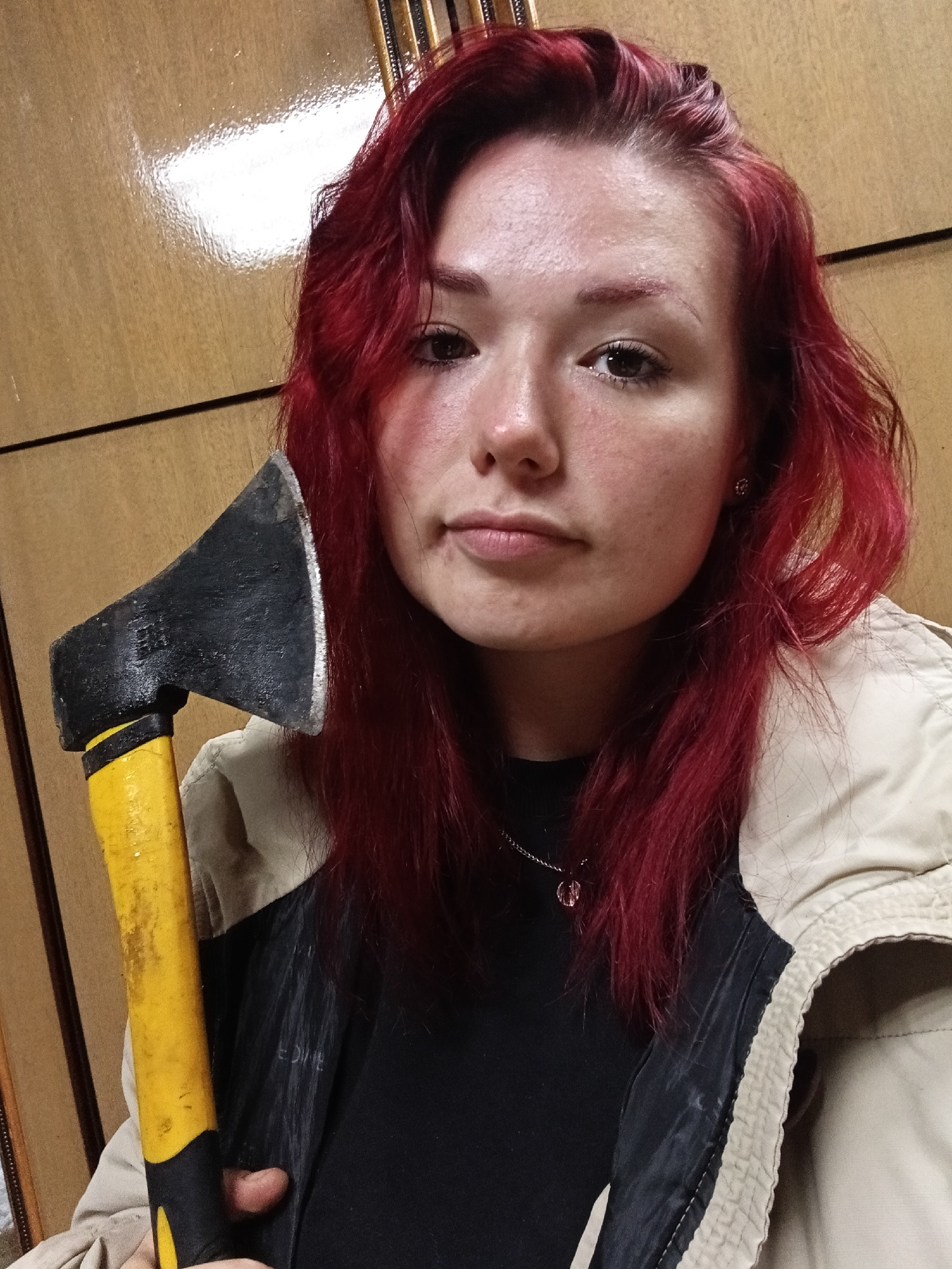 A young woman with bright red hair is holding an axe in the foreground. She is looking at the camera with a neutral expression. She is wearing a black t-shirt, a beige jacket, and a silver necklace with a pink pendant. The axe has a yellow handle and a black head. The background is a wooden wall. The image appears to be taken indoors, in an unknown urban location.