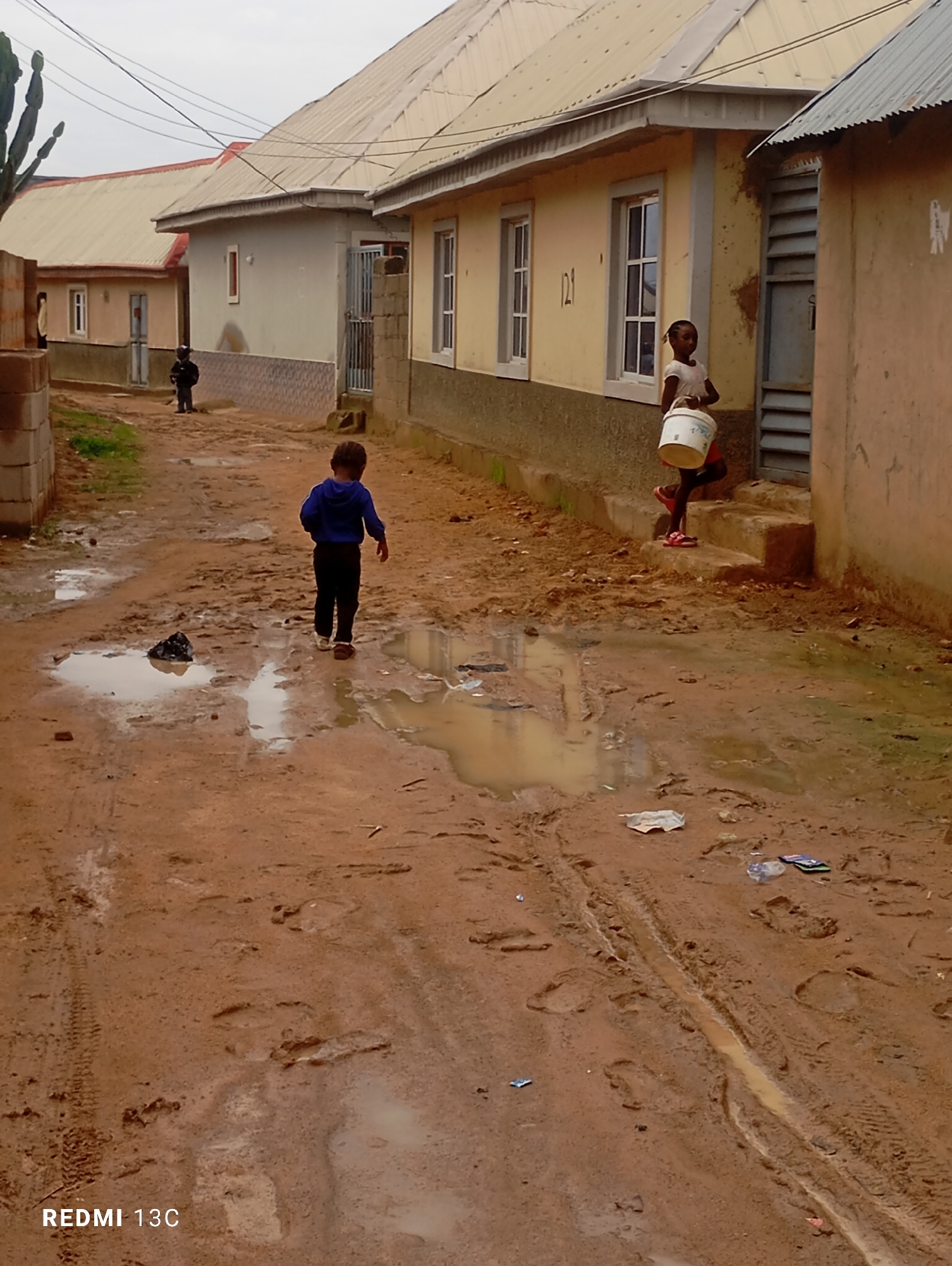 In Jos, Nigeria, after a heavy rain, a muddy street in front of residential buildings is visible. The street is covered in puddles and tire tracks. A young boy in a blue sweatshirt and dark pants walks away from the camera, his back to the viewer. He is walking through a puddle. Further down the street, a young girl wearing a white t-shirt and red shorts carries a white bucket. She is walking up a set of concrete stairs. One building has a yellow facade with white-trimmed windows, and another building has a pale grey facade. Overhead, electrical wires are strung across the sky. The image appears to have been taken from a slightly elevated position, looking down the street.