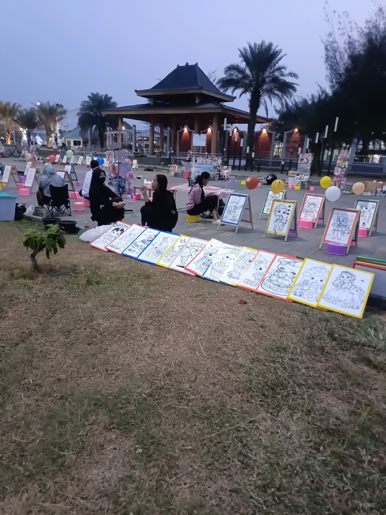 The image depicts an outdoor scene at dusk, likely in the city of Lubuklinggau, Indonesia. In the foreground, a row of coloring pages, some with cartoon characters and designs like Cinderella and Hello Kitty, are laid out on the grass. Behind the coloring pages, several people are seated on the ground or in chairs, appearing to be engaged in an activity, possibly drawing or coloring. Some individuals are wearing face masks. In the background, a traditional-style pavilion with a dark, multi-tiered roof is visible, surrounded by palm trees and other vegetation. There are also lights illuminating the area, suggesting a public space or event. Balloons are scattered around the scene. The ground is covered with grass and a paved area. The overall atmosphere suggests a community gathering or a street fair.
