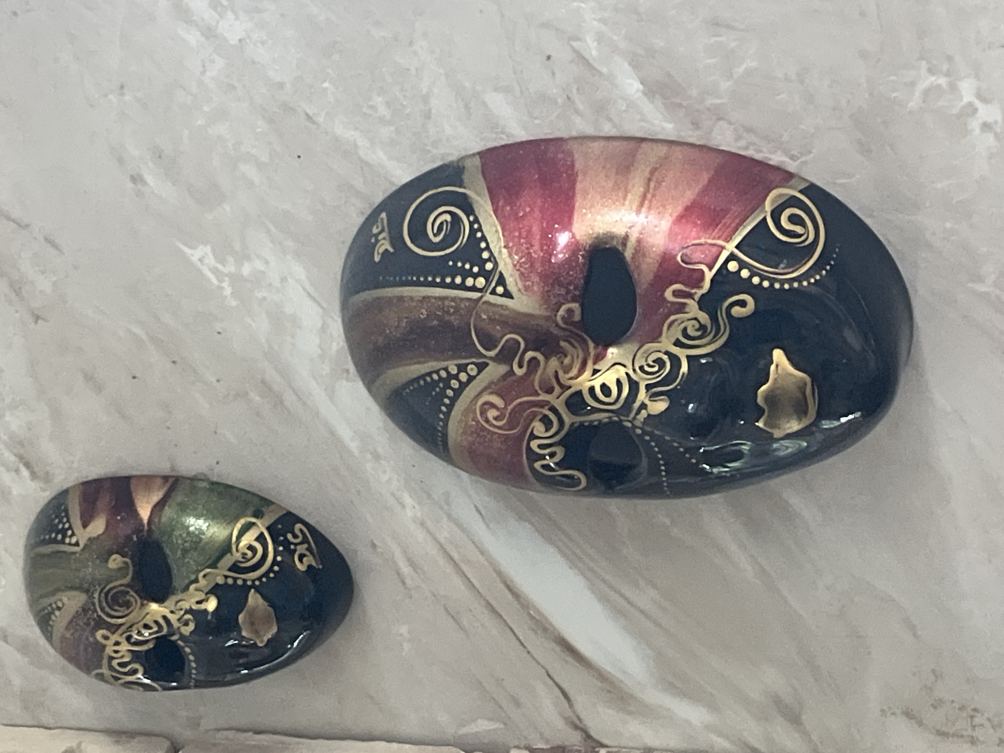 The image shows two decorative masks mounted on a textured wall. The masks are oval-shaped with eyeholes and are painted in dark blue, red, gold, and green. They feature ornate gold swirls and dotted patterns. The mask in the foreground is larger and more detailed, with a gold mouth. The smaller mask in the background is partially visible. The wall appears to be made of a light-colored, possibly stone or textured plaster material. The lighting suggests an indoor setting. There is no indication of the city of Vladimirovka, Russia in the image.