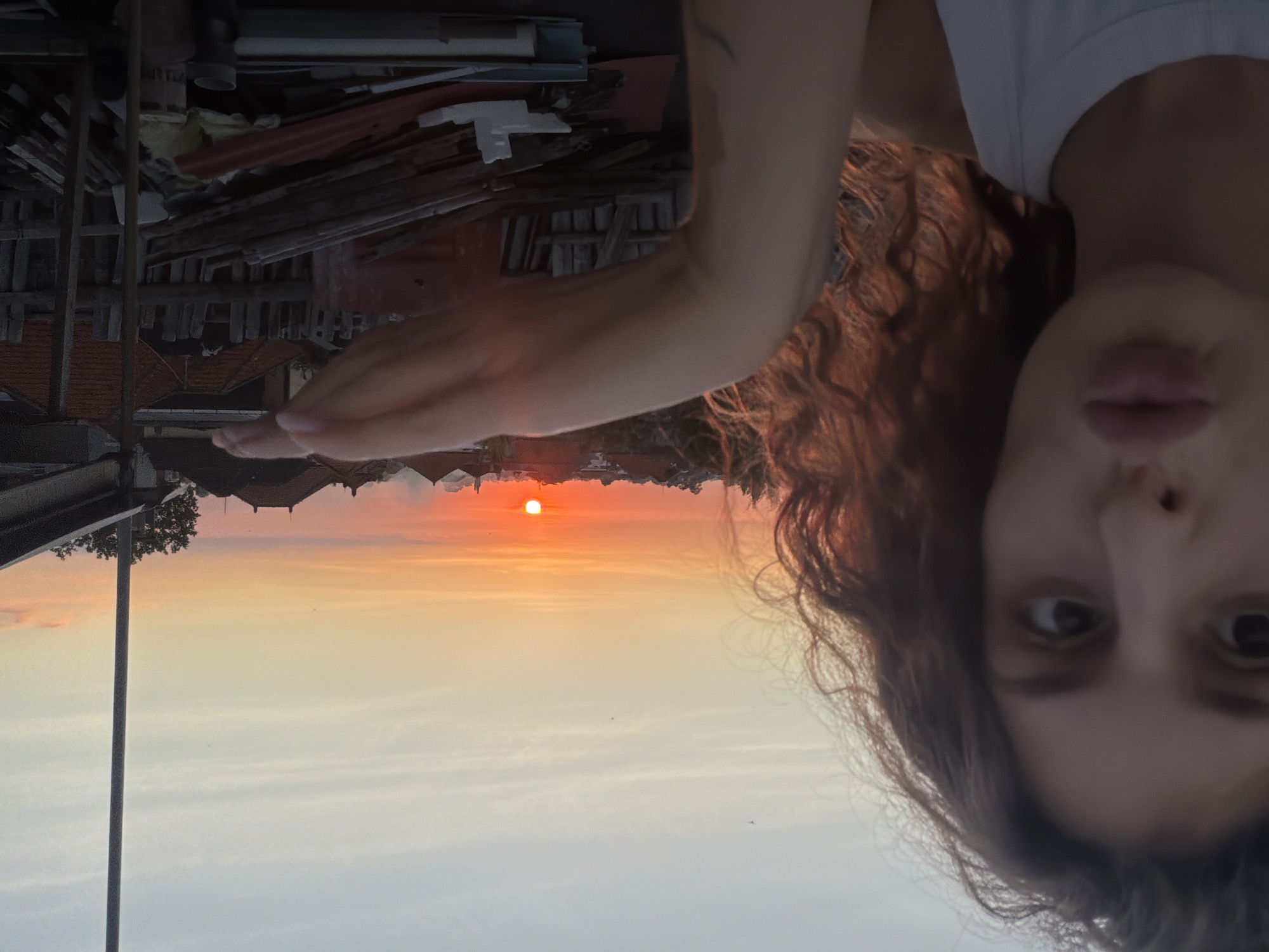 An upside-down, medium shot features a woman with curly brown hair, a hand outstretched towards a sunset, and a background view of North Kuta, Indonesia. The buildings have red roofs. There is a metal post on the left. The sky is a gradient of pale orange and white around the sun. The woman’s face is partially in focus. Debris, including lumber and roofing materials, is visible above the roofs.