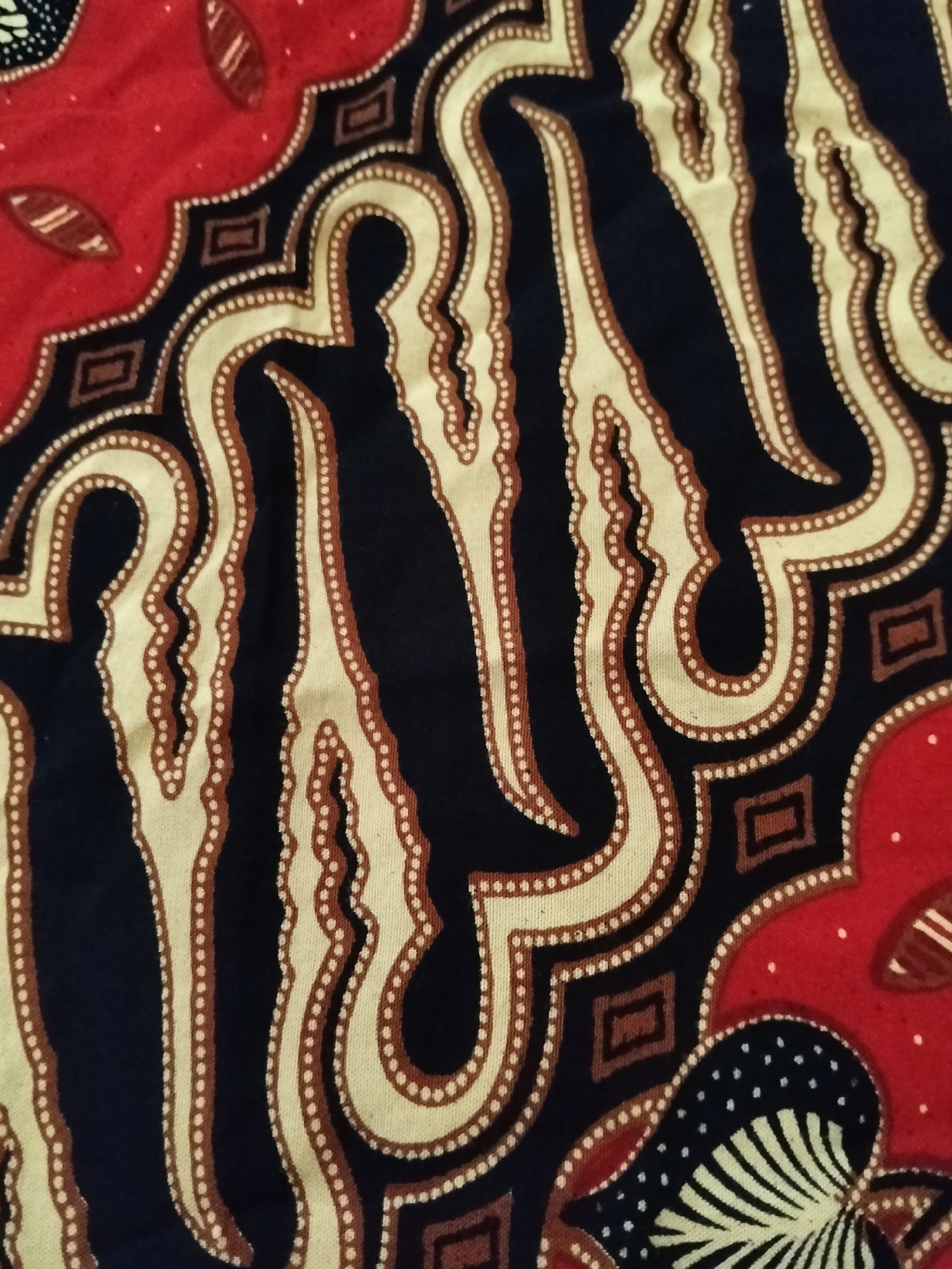 The image is a close-up, high-angle shot of a piece of red, black, and beige batik fabric. The fabric features an intricate pattern of organic, wavy lines and geometric shapes, with a dotted border detail. There are no people, actions, or interactions depicted. The environment is not discernible beyond the texture and pattern of the fabric itself. No information about the location in Gempolan, Indonesia is present in the image.