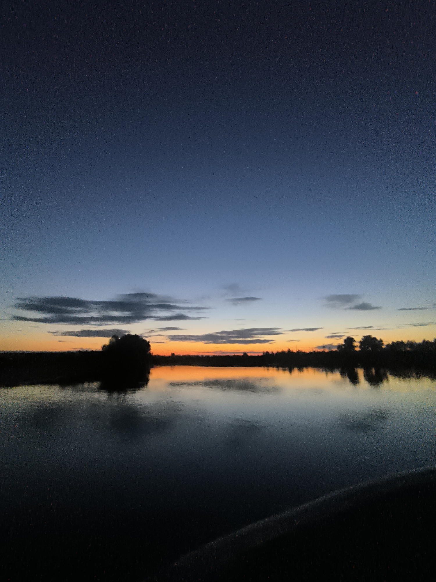 This is a vertical, full-shot, eye-level, outdoor photograph taken at dusk in Zagotzerno, Russia. The scene depicts a tranquil body of water reflecting the colorful twilight sky. Silhouetted trees and land form a dark line along the horizon, with a large, dark mass of trees on the left and smaller clusters of trees scattered to the right. The sky transitions from a deep blue at the top to a vibrant orange and yellow band just above the horizon, with faint, wispy clouds present. The water is dark and still, mirroring the sky and the land. The foreground is dominated by the dark water, with a hint of a dark, sloped surface in the lower right corner, possibly a bank or a road. There are no people or animals visible in the image.