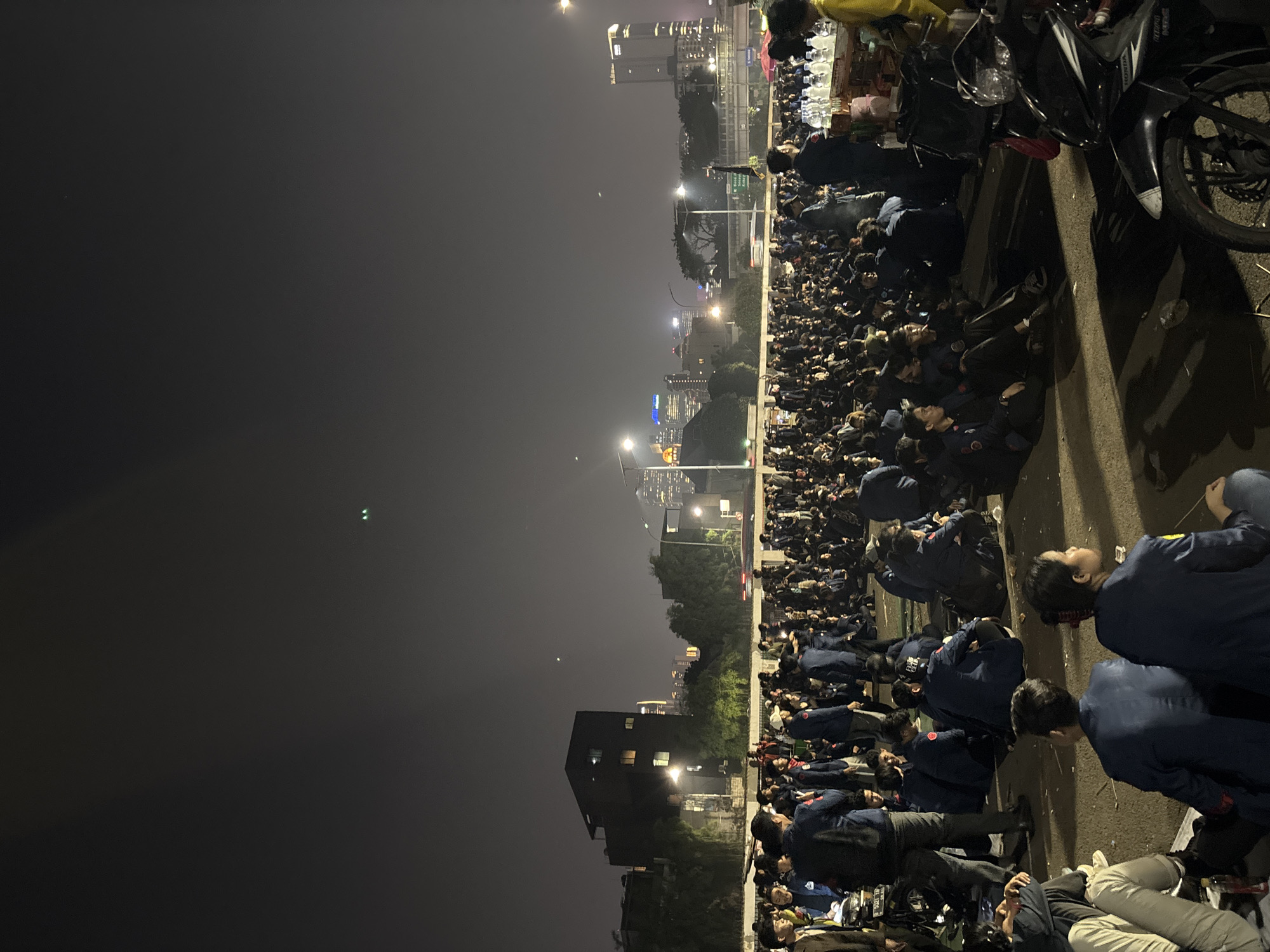The image depicts a large gathering of people, predominantly young adults dressed in matching dark blue uniforms with yellow accents, sitting on the ground in what appears to be an urban outdoor setting at night. The scene is illuminated by streetlights and the lights of distant city buildings, suggesting a location within a developed urban area. In the background, several tall buildings and city infrastructure, including overpasses and streetlights, are visible, indicating a city environment. The presence of motorcycles parked on the right side of the frame further suggests a public urban space. The overall atmosphere appears to be one of a planned assembly or event, with the individuals seated in an organized manner. The specific location is identified as being in the city of Central Jakarta, Indonesia.