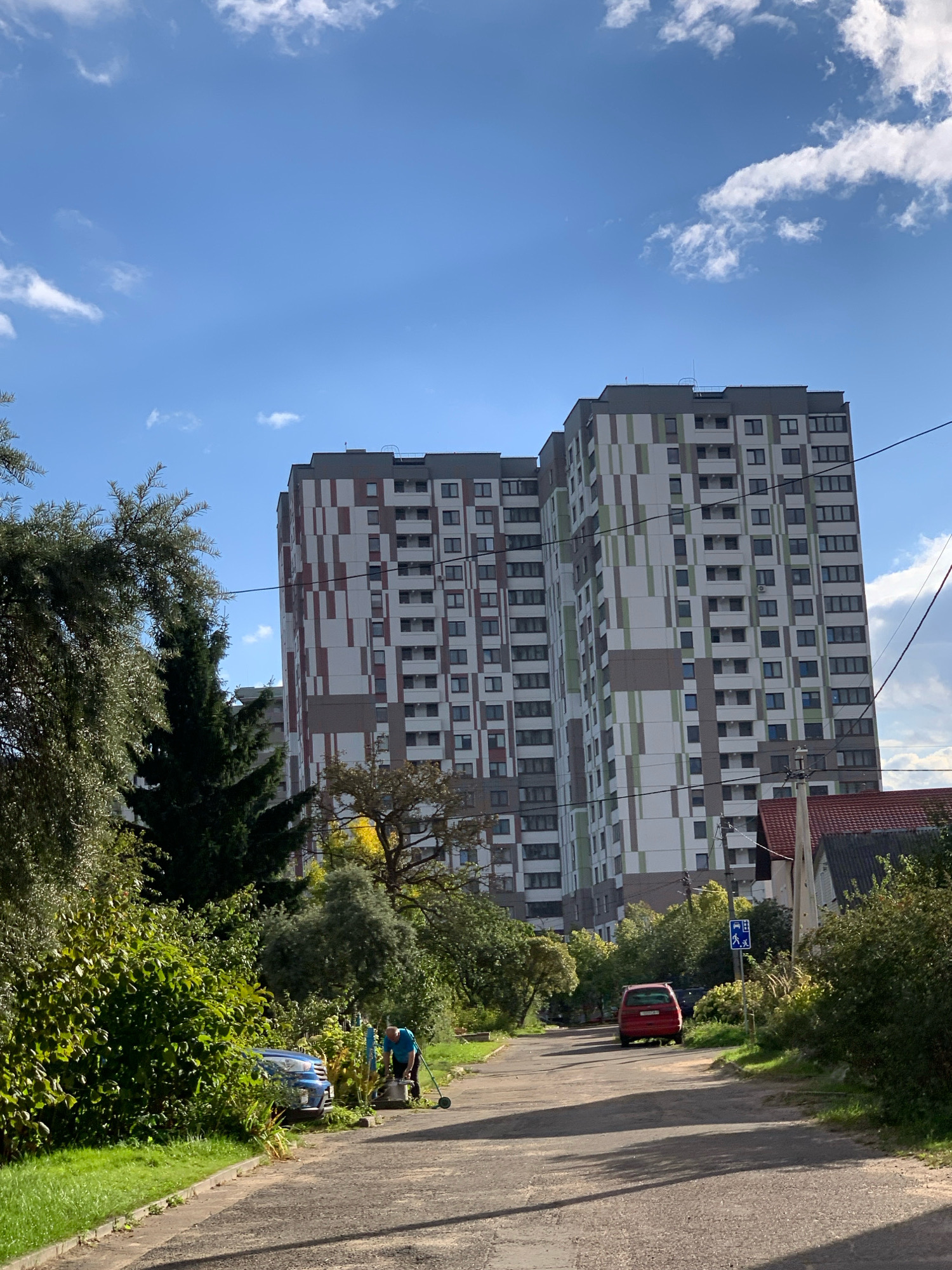 The image depicts a sunny day in Minsk, Belarus, with a clear blue sky dotted with white clouds. In the background, two modern, multi-story apartment buildings stand tall, characterized by their distinctive facades featuring a geometric pattern of white, gray, green, and reddish-brown rectangles. A paved road leads towards the buildings, with a red car parked on the right side of the road. On the left side of the road, dense green foliage and trees partially obscure a blue car, and a person is seen operating a lawnmower. A pedestrian crossing sign is visible on the right side of the road, indicating a designated crossing area. The overall scene suggests a residential neighborhood on a pleasant day.