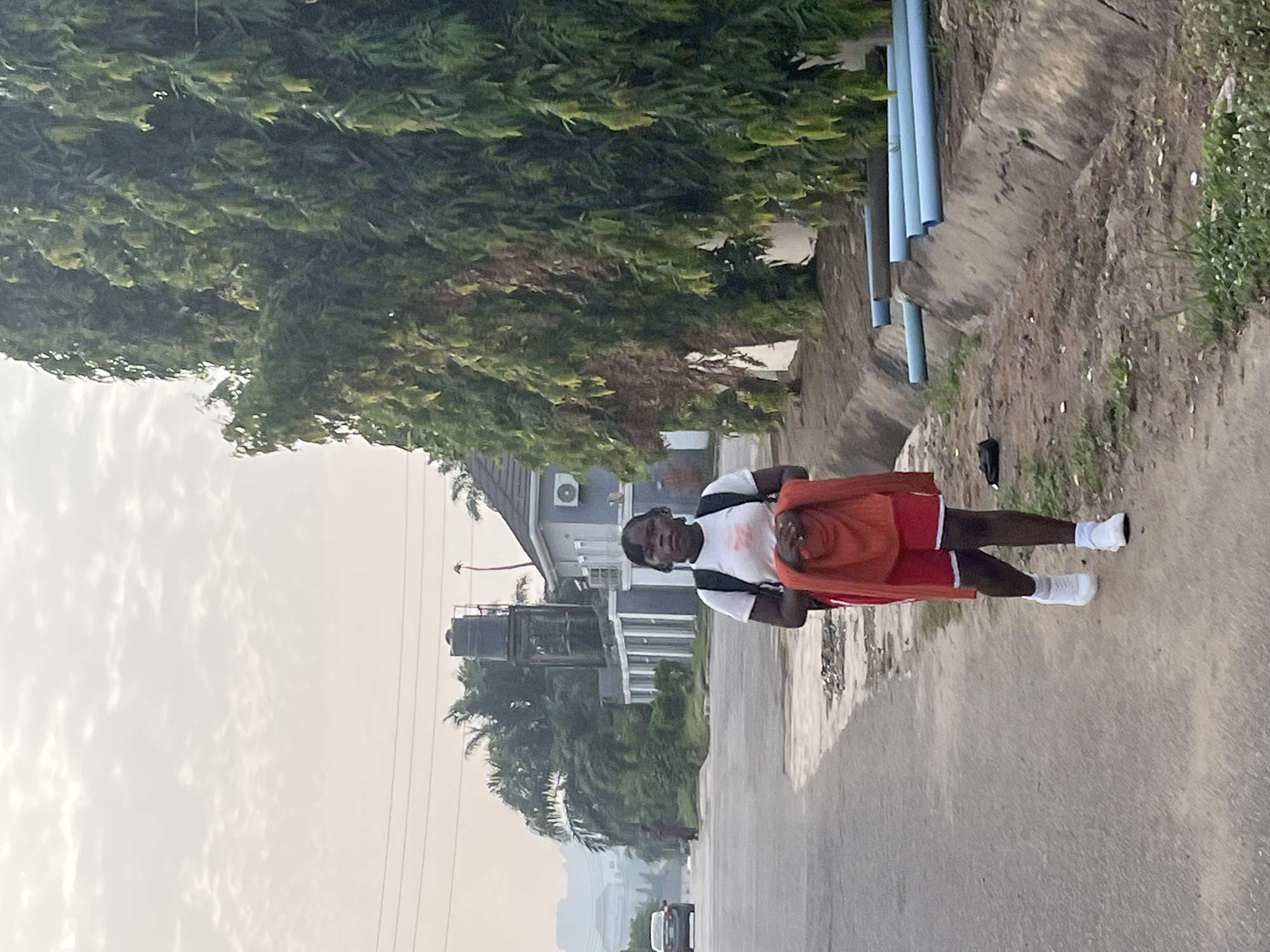 A man stands on an asphalt road in a residential area. He wears a white t-shirt, red shorts, and white sneakers, and carries a folded orange garment. Behind him is a house with a water tank and tall green trees. Above him, there are blue pipes on a concrete structure. The sky is overcast. Cars are visible in the distance. The location appears to be a street in Abuja, Nigeria, based on the architecture and vegetation.