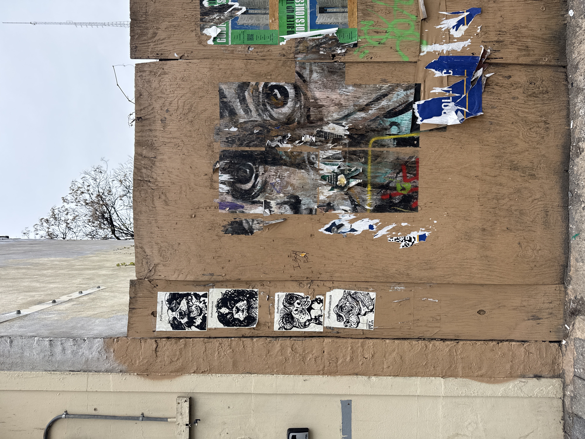 The image depicts a plywood wall covered with torn posters and stickers in an urban environment. The wall is composed of horizontal wooden planks, painted brown. Above the wall is a building corner painted light grey. The remnants of posters are glued to the top planks of the wooden structure. A large portion of one poster appears to depict a tiger’s eyes, which are dark with some visible brown. Below the eye imagery, more posters are torn. On the bottom of the wooden wall, there are four similar-looking, black and white stickers displaying illustrations of dogs. Below the stickers, the wall is painted brown, and then a lighter beige color. Attached to the bottom wall is a gray pipe. A tree and antenna are seen above the building on the left side. The location is the city of New York, United States of America. No people are visible in the image. The overall impression is one of urban decay and street art.