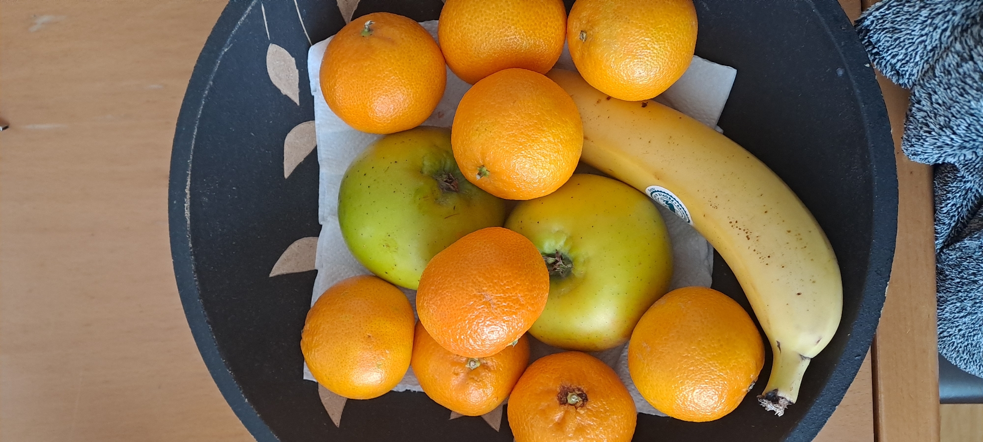 In the city of Castlebar, Ireland, a black, textured bowl contains an assortment of fruit. The bowl sits on a wooden surface, and a grey and white patterned textile is visible at the bottom edge of the frame. Inside the bowl, on top of a white paper towel, are two green apples, a bunch of tangerines, and a single yellow banana. The tangerines are scattered around the apples, and the banana curves across the bottom of the bowl, partially overlapping the fruit. One tangerine has a sticker on it. The lighting suggests an indoor setting, likely a kitchen or dining area.