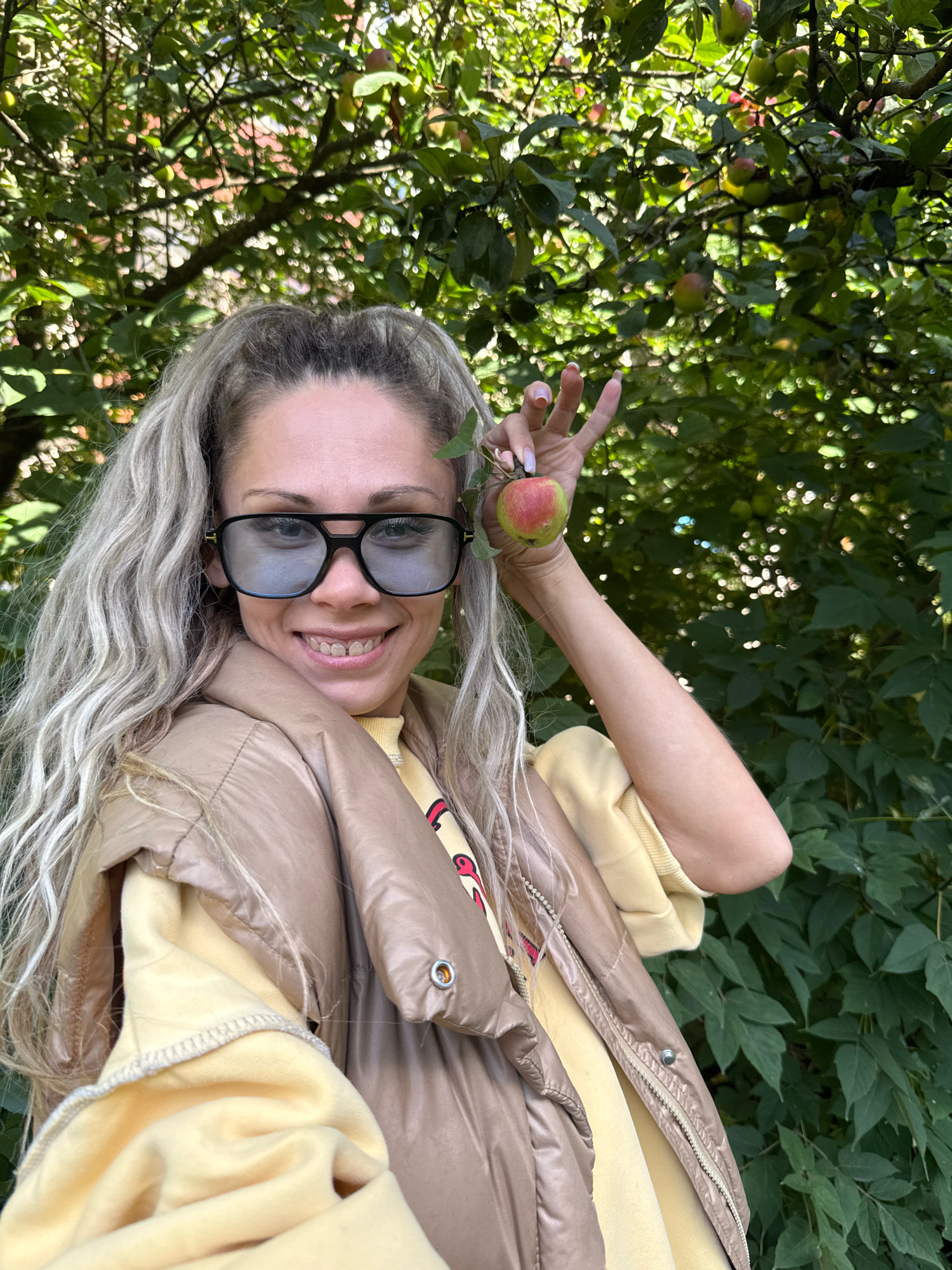 A medium shot shows a woman with long, wavy blonde hair and fair skin posing outdoors in front of a tree with green leaves and apples. She is wearing large, dark-rimmed sunglasses, a tan puffer vest over a yellow sweatshirt, and a light pink nail polish on her fingernails. The woman is holding a small, red and green apple in her right hand, raised near her face. The background is filled with the green foliage of the tree and dappled sunlight. The woman is smiling directly at the camera. The location is identified as Rusanovo, Russia.
