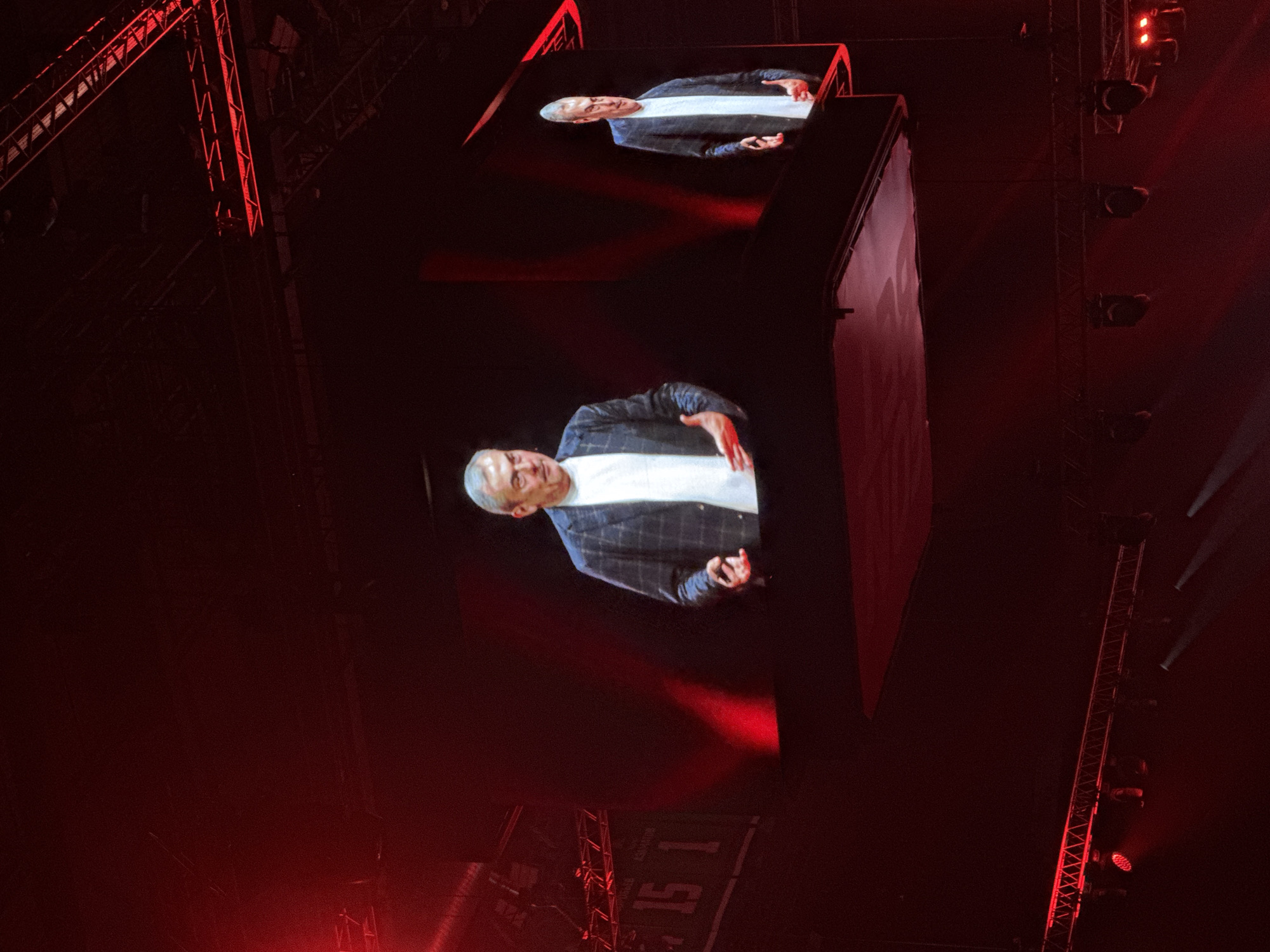 The image depicts a stage with a large screen displaying a live feed of a man in a checkered jacket and white shirt. The man is gesturing with his hands. To the left of the screen, a scoreboard is visible with the number "15 1" on it. The environment is predominantly dark, with red lighting illuminating the stage and parts of the audience area. The background shows a complex stage rigging system with lights. The scene appears to be a presentation or a performance. It is not possible to determine the specific location in Ufa, Russia, from the image alone.