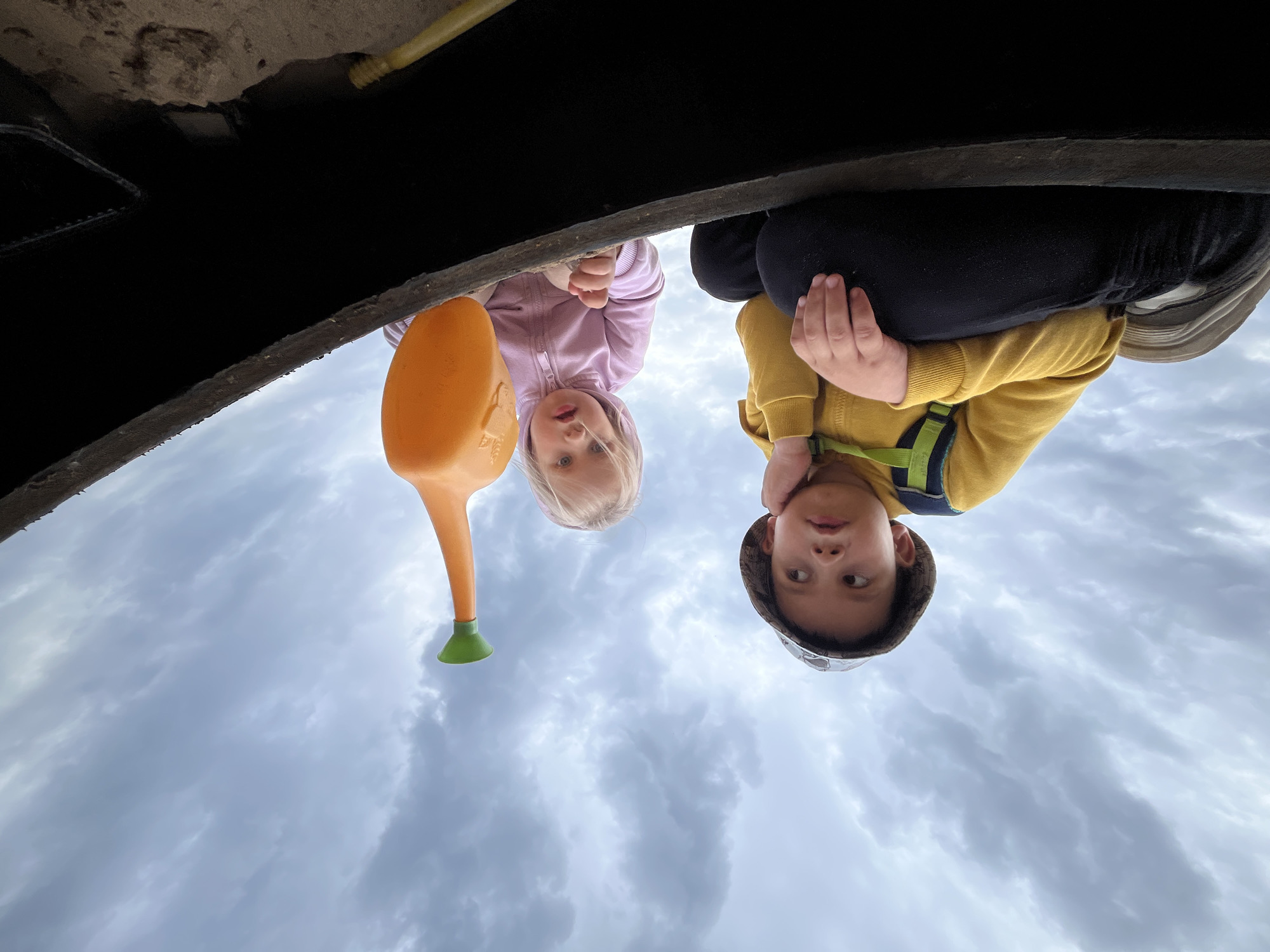 Two children and a watering can are seen from an unusual perspective, looking down at the camera. A toddler wearing a pink jacket stands to the left, next to an orange watering can with a green spout. A young boy wearing a yellow jacket and a camouflage-patterned hat sits to the right. They are positioned under a dark, elevated structure, with a view of a cloudy sky filling the background. The specific location in Chaikovsky, Russia, cannot be determined from the image.