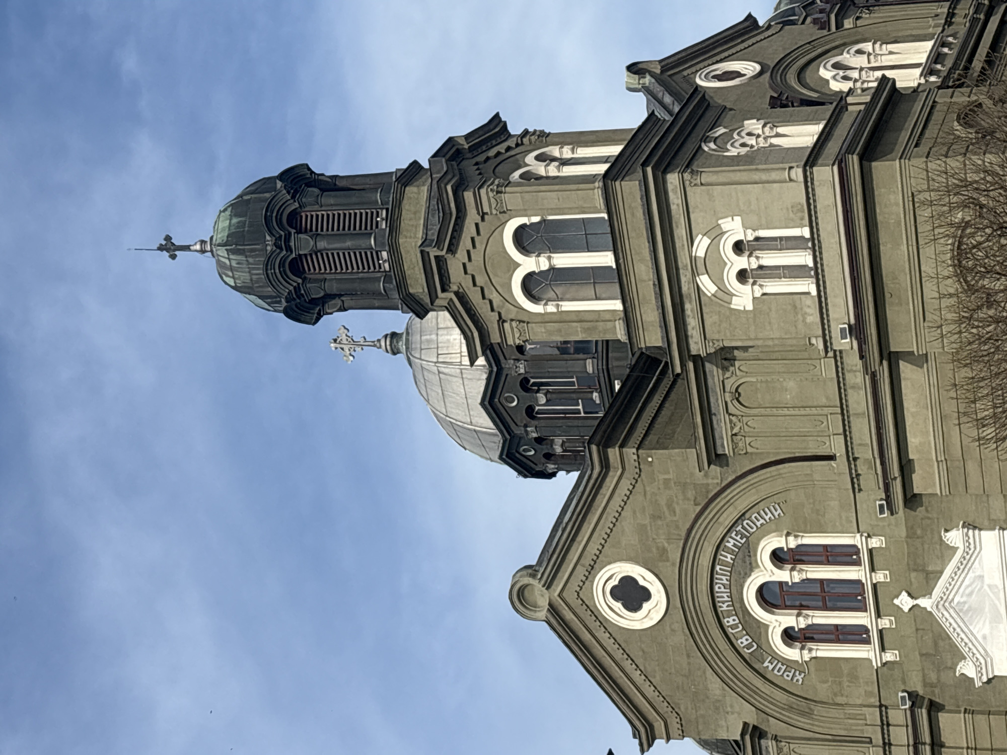This image captures a low-angle view of the Church of Saints Cyril and Methodius in Burgas, Bulgaria, set against a bright blue sky with light, wispy clouds. The building is a grand, ornate structure, primarily dark grey with striking white decorative elements around its windows and architectural features.
A prominent bell tower dominates the left side of the frame, featuring a dark, ribbed dome culminating in a tall spire topped with an Orthodox cross. Below this, a second, lighter-colored dome is partially visible. The church's facade is rich in detail, showcasing multiple arched windows, some with white, triple-lobed frames, and small circular or clover-shaped apertures.
Above a large set of triple-arched windows, a significant inscription in Cyrillic script arcs across the facade. The text reads: "ХРАМ СВ. СВ. КИРИЛ И МЕТОДИИ", which translates to "CHURCH OF SAINTS CYRIL AND METHODIUS."
On the far right, the bare branches of a tree are visible, suggesting the season might be late autumn, winter, or early spring. The clear sky and distinct shadows indicate it is daytime, likely a bright and sunny morning or afternoon. No people or activities are present, allowing full focus on the architectural beauty and solemnity of the religious edifice.