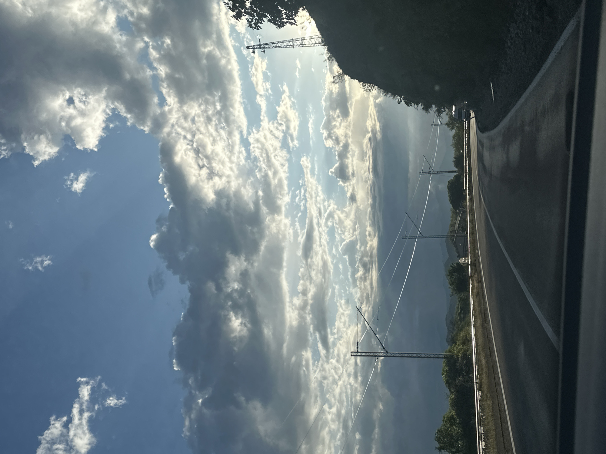 The image captures a dynamic landscape view, likely taken from a moving vehicle, showcasing a road and an expansive, dramatic sky near Vranjina, Montenegro.
The sky is a prominent feature, filled with large, voluminous cumulus clouds. Sunlight breaks through gaps in the cloud cover, illuminating some clouds brightly and creating strong highlights, while other areas of the sky and clouds appear darker and more shadowed, suggesting varying cloud thickness and light conditions. Patches of clear blue sky are visible in various parts of the frame.
On the right side, a segment of a paved road extends into the distance, marked by a solid white line at its edge. The road surface appears somewhat reflective, possibly indicating dampness from recent rain. Running parallel to the road, a series of metal lattice poles support an overhead electrical or catenary system, with numerous wires stretching across the middle ground.
To the left of the road, a strip of lush green vegetation, consisting of trees and bushes, borders the roadside. In the farther distance, dark, undulating mountains or hills are visible against the horizon. A small white road marker or sign can be seen beside the road further along. The lighting suggests either late afternoon or early morning, with the sun low in the sky, creating a strong interplay of light and shadow. There are no people or discernible text visible in the image.