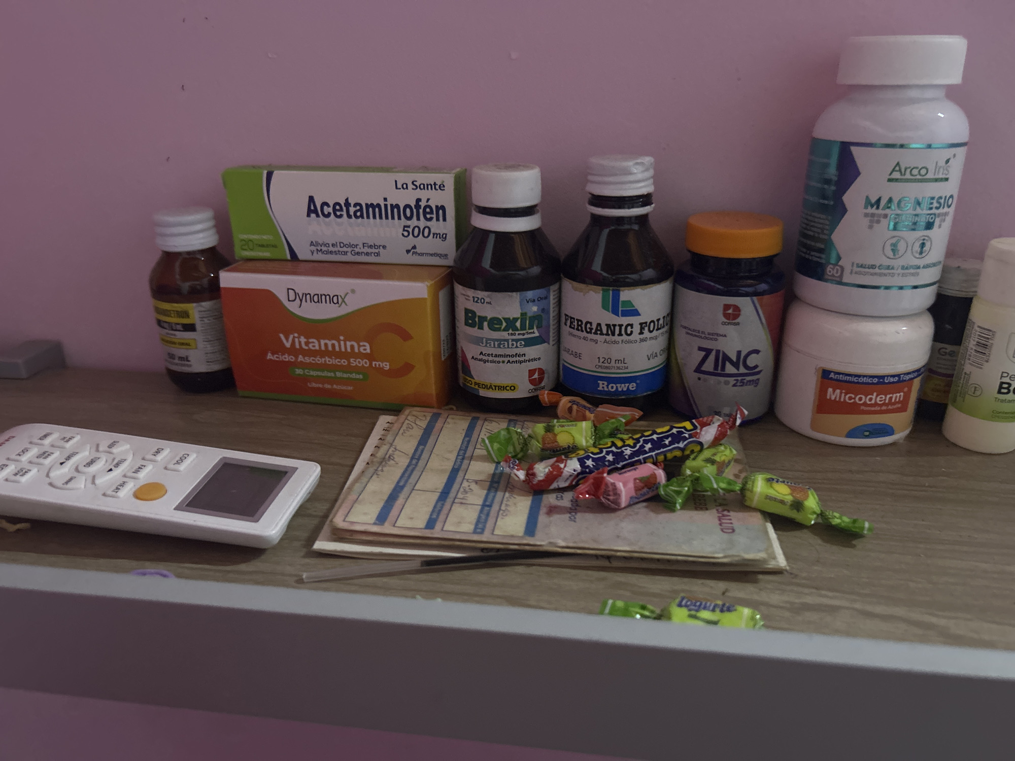 The image presents a collection of medicinal products and other objects arranged on a wooden shelf or surface. Dominating the background are several boxes and bottles of medication, including Acetaminofen, Dynamax vitamin C, Brexin syrup, Ferganic Folic, Zinc tablets, Arco Ins magnesio, and Micoderm antifungal cream. A small bottle with an illegible label is also visible. In the foreground, to the left, there is a white television remote control. Adjacent to it is a stack of papers or a small notebook, partially covered with candies of various colors and flavors. The background is a plain pink wall. The image does not contain sufficient information to determine a specific location within San Juan de Los Morros, Venezuela.