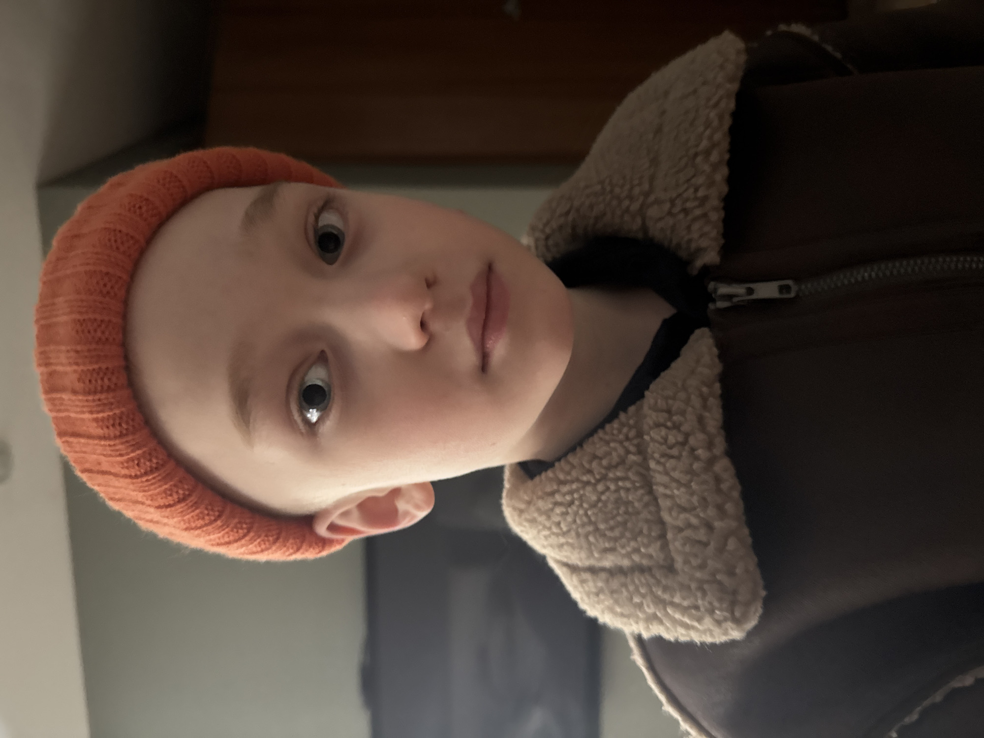 The image displays a close-up, frontal view of a young individual, likely a child or pre-teen, situated indoors in Votkinsk, Russia. The individual has fair skin, light-colored eyes, a small nose, and closed lips, maintaining a neutral expression while looking directly at the camera. They are wearing an orange, ribbed knit beanie. The upper body is clad in a dark brown jacket featuring a distinct light brown, textured (faux shearling or sherpa-style) collar. A silver-colored zipper is visible down the front of the jacket, partially unzipped over a dark undershirt. The background is out of focus; a dark, rectangular object, potentially a television screen, is partially visible to the upper left. The wall to the upper left appears light grey, while the right side of the background shows a darker, brownish surface, possibly a door or a different wall.