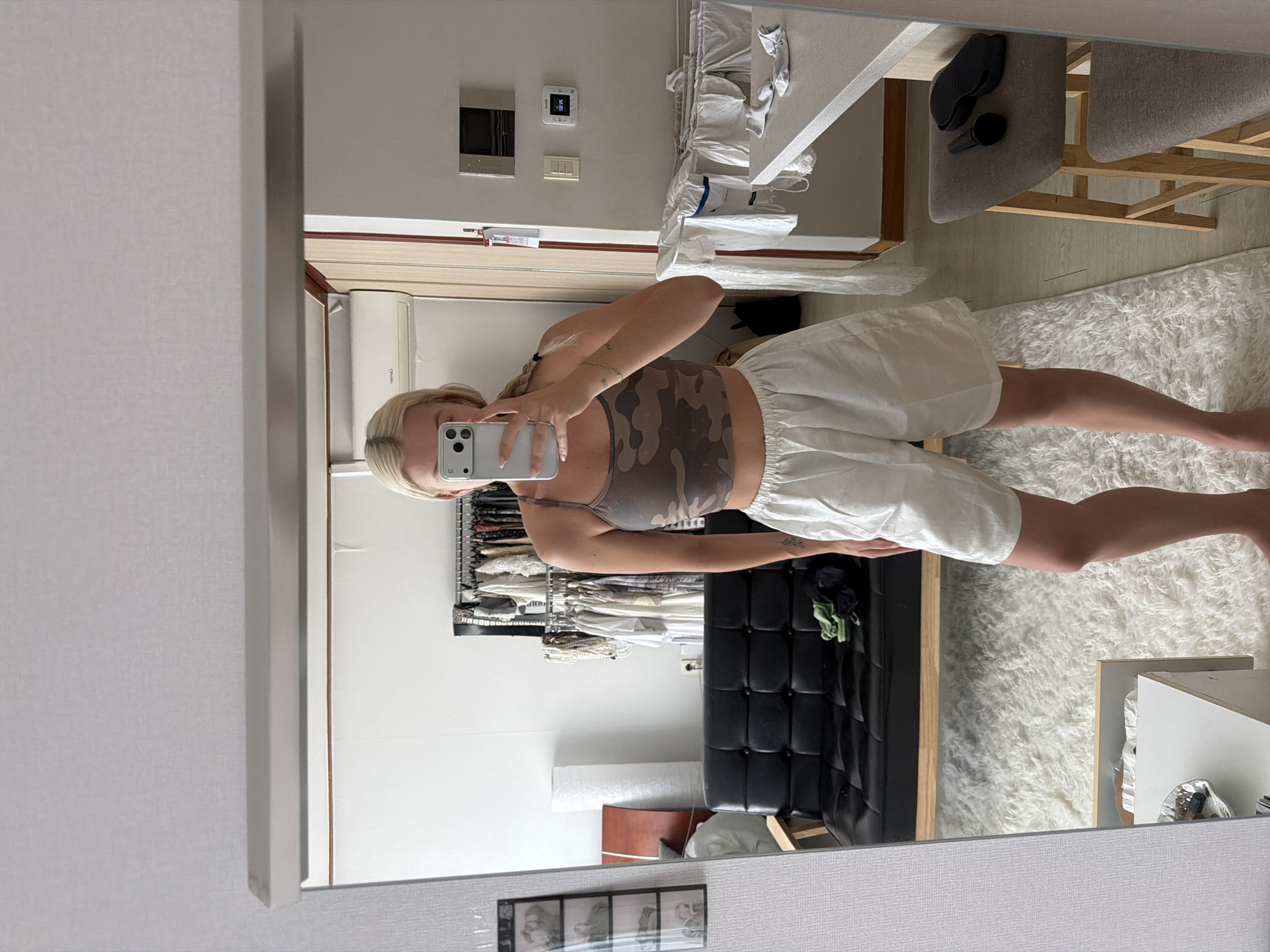 A mirror selfie depicts a woman with blonde hair, wearing a camouflage print tank top and white shorts. She is holding a smartphone in her right hand, seemingly capturing the image. The backdrop includes a wardrobe, a black leather couch, and white fluffy rug. The setting appears to be an indoor space, likely a residential area. It is not possible to determine the specific location in Seoul or confirm that this image was taken in Seoul, South Korea based solely on visual information.