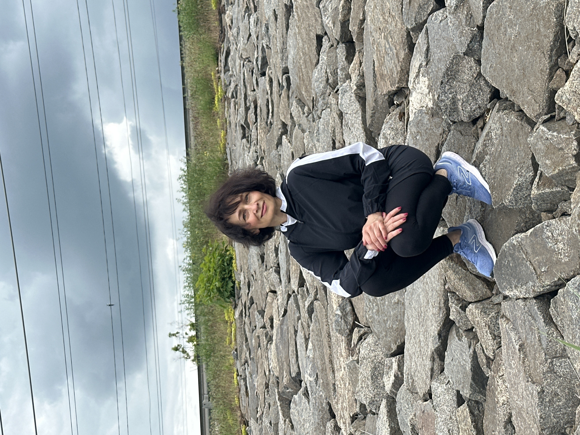 A middle-aged woman with short dark hair is crouching on a rocky surface. She is wearing a black track jacket with white stripes on the sleeves, black leggings, and light blue athletic shoes with a white "N" logo. Her hands are clasped around her legs, and she is looking toward the viewer. The background consists of grey rocks of varying sizes, stretching behind the woman. To the left of the rocks, there is a line of green vegetation. Above the vegetation, there are electrical power lines visible against a cloudy sky. The location appears to be an embankment or a rocky area next to a roadside with a grassy verge. The city is unknown.