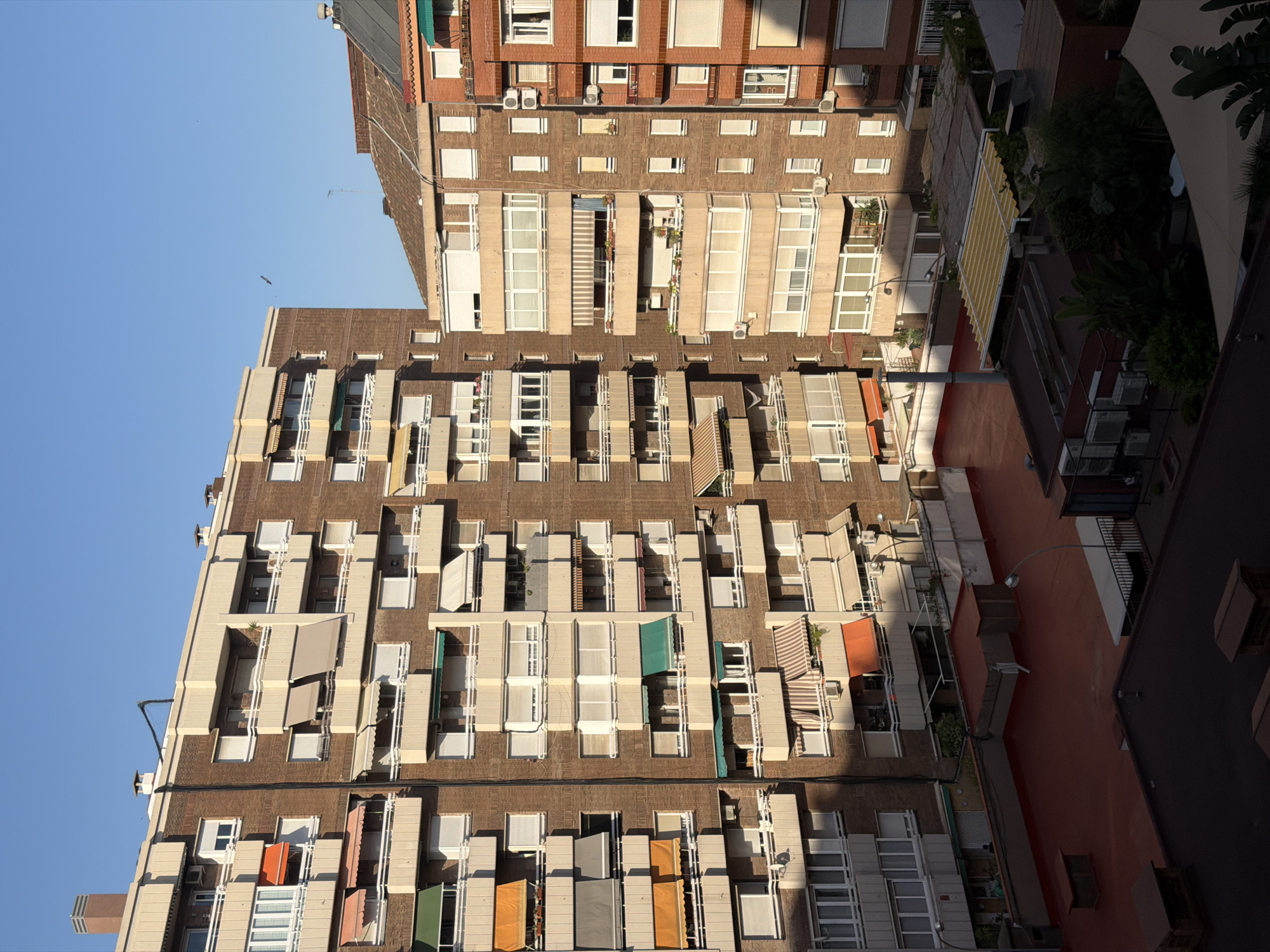 This is a high-angle, vertical shot of apartment buildings in Barcelona, Spain, during the daytime. The sun is casting long shadows, suggesting it is either early morning or late afternoon. The buildings are constructed primarily of brick with lighter colored balconies and window frames. Many of the balconies are equipped with retractable awnings in various colors, including beige, orange, yellow, and green, indicating a sunny climate.
The scene depicts the architecture of a residential urban area, with multiple stories of apartments visible. Some balconies have plants, adding touches of greenery to the brick structures. Air conditioning units are visible on some of the buildings. The sky is clear and blue. The image is taken from a vantage point looking down onto the buildings, possibly from another building or a higher floor. No people are clearly visible, and the focus is on the built environment. The overall impression is one of a typical European city dwelling scene, with a focus on the interplay of light and shadow on the building facades.