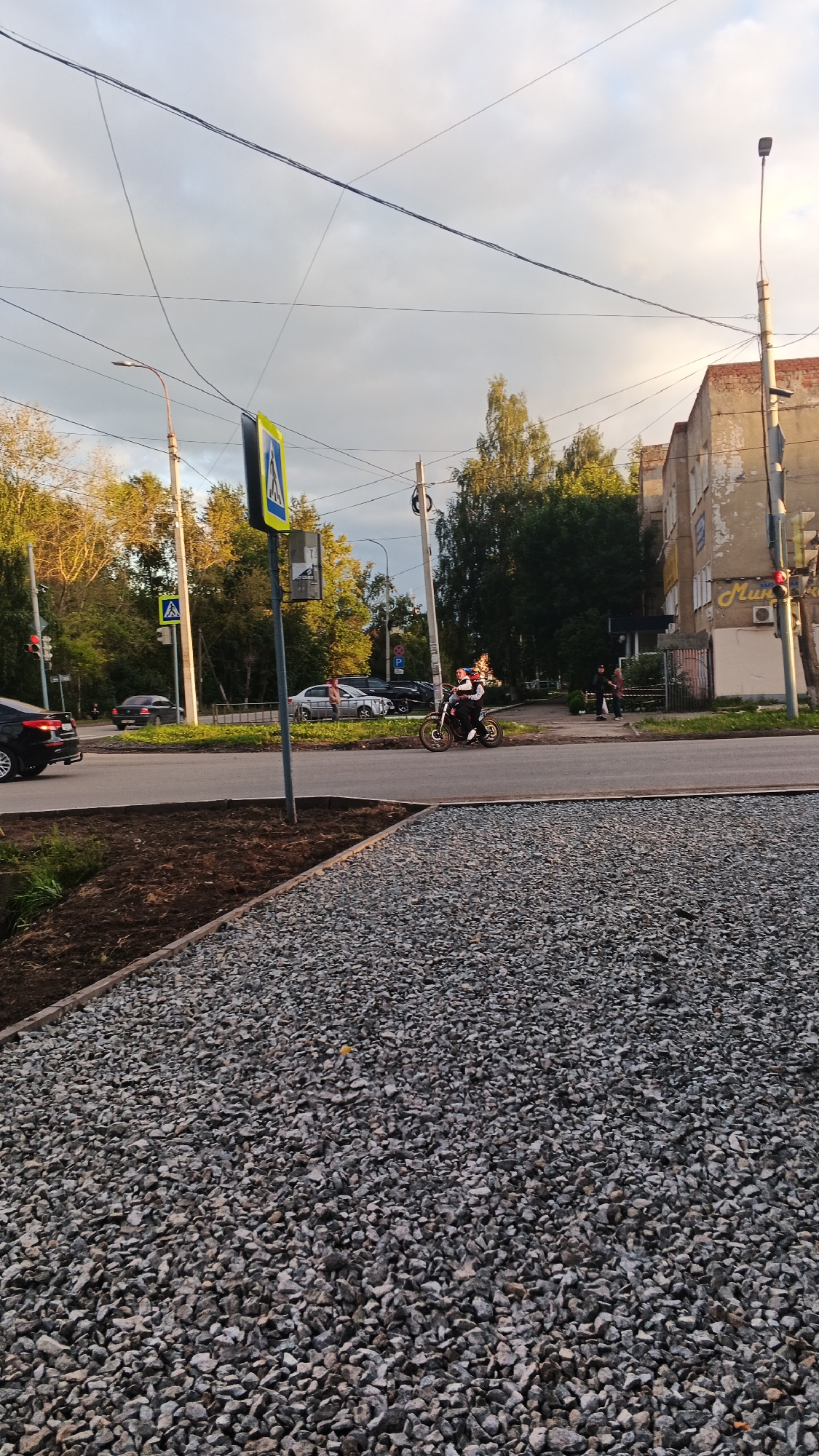 The image captures a scene on a city street in Izhevsk, Russia, likely during the late afternoon or early evening, given the lighting. In the foreground, a patch of rough gravel and dark soil borders the paved road. The road itself is a multi-lane thoroughfare with visible lane markings.
Several vehicles are present: a black car is seen in the left portion of the frame, and other cars, including a silver sedan and a dark SUV, are stopped or moving in the background. A pedestrian crossing is indicated by a yellow and blue sign on a pole.
The central focus of activity is a person on a motorcycle, wearing a white, red, and black outfit. The individual is positioned on the road, appearing to be either stationary or moving slowly. Other individuals are also visible in the background, near a building.
The background features apartment buildings with weathered facades, indicative of urban development. Trees and streetlights are interspersed along the roadside. Overhead, a network of electrical wires crisscrosses the sky, which is partly cloudy with a mix of light and shadow. The overall impression is a typical urban streetscape.