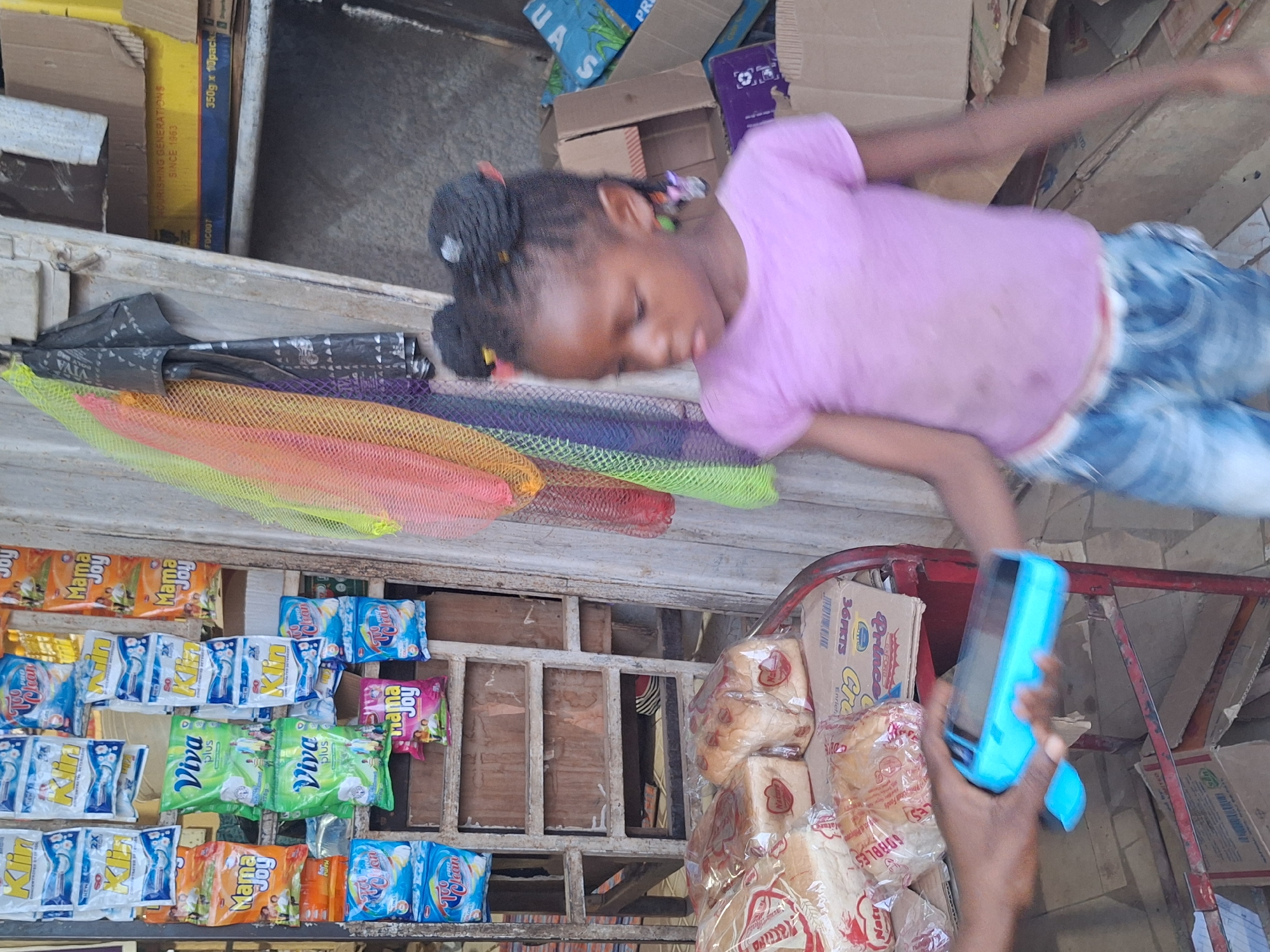 The image depicts a candid scene within a market stall or small shop in Sabon Tasha, Nigeria. A young girl, appearing to be a child or pre-teen, with dark skin and braided hair styled in two buns, is resting or sleeping on a counter or ledge. She is dressed in a light purple t-shirt and blue jeans.
The surroundings are filled with various goods. To the left, numerous packets of detergents and soaps are displayed, including brands like "Klin," "Viva plus," "Mama Joy," and "Too Clean." Above these, colorful mesh sponges in red, orange, yellow, green, and purple are visible. To the right of the girl, several loaves of bread, some labeled "Nakar's" and "GOBBLES," are stacked in clear plastic bags.
The background shows multiple cardboard boxes, with readable text such as "OXFORD," "Prince," "NOURISHING GENERATIONS SINCE 1963," "350g x 10pack," and "FDC007." Part of an adult's hand, holding a bright blue mobile phone, is visible on the far right, near a red metal cart. The lighting indicates it is daytime. The scene captures a moment of rest amidst the daily operations of a local shop.